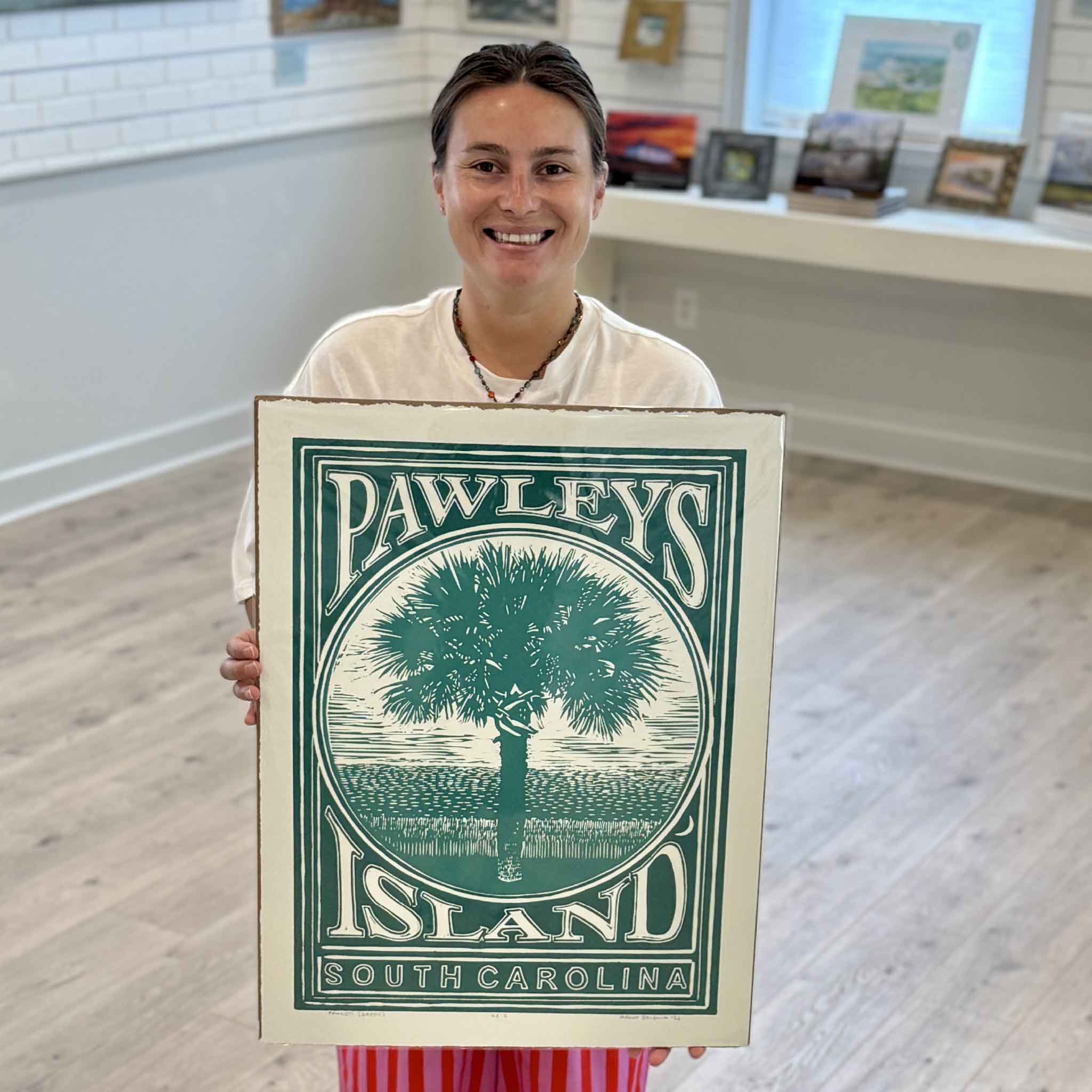 Kyra of Gray Man Gallery holding the green edition of Aaron Baldwin's Pawleys Island linocut featuring a palmetto tree and coastal marsh landscape.
