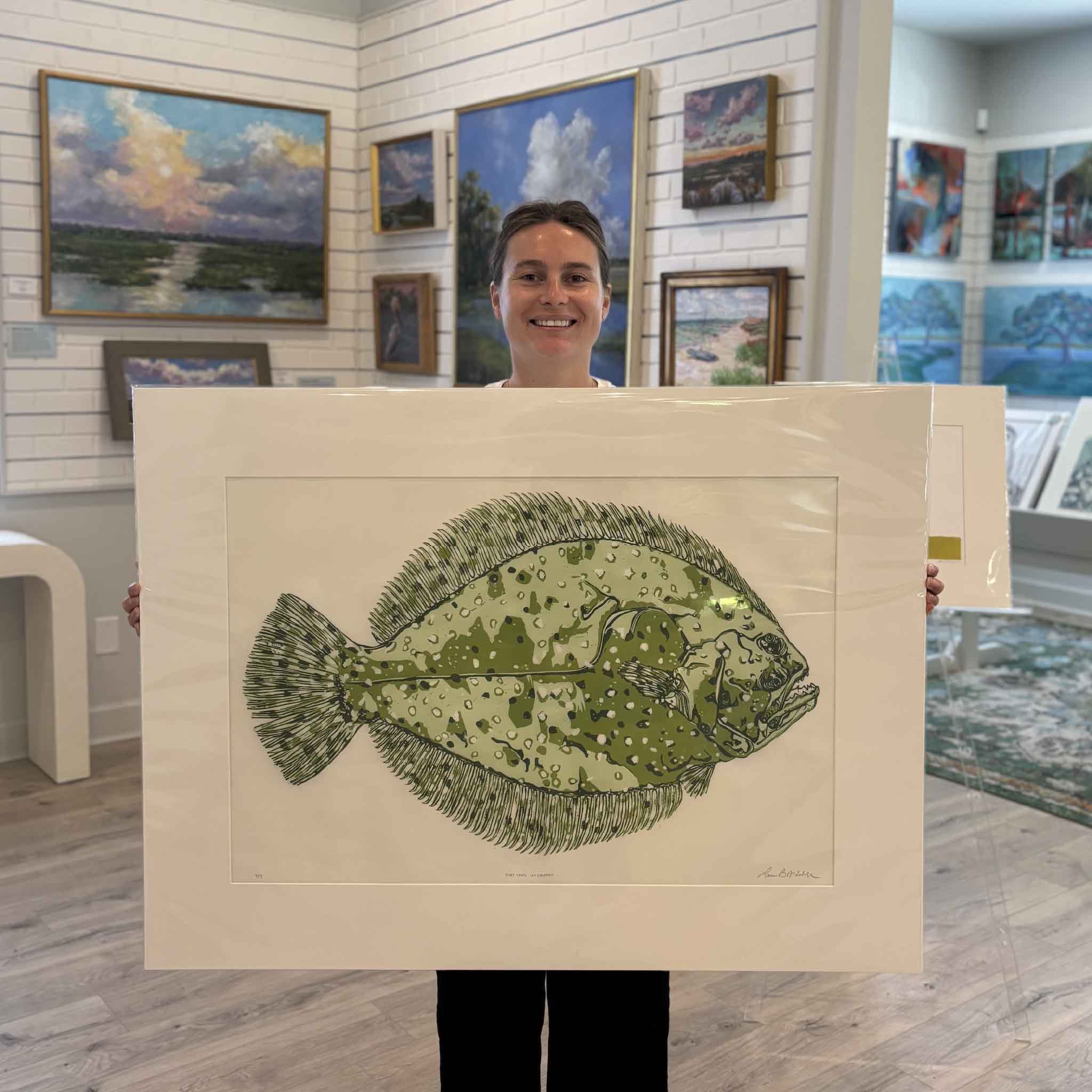 Kyra holds Flat Fred in Green, an original signed reduction linocut by Laurie Brownell McIntosh featuring her iconic Lowcountry flounder. This professionally matted, hand-pulled original artwork is available exclusively at Gray Man Gallery in Pawleys Island, South Carolina.