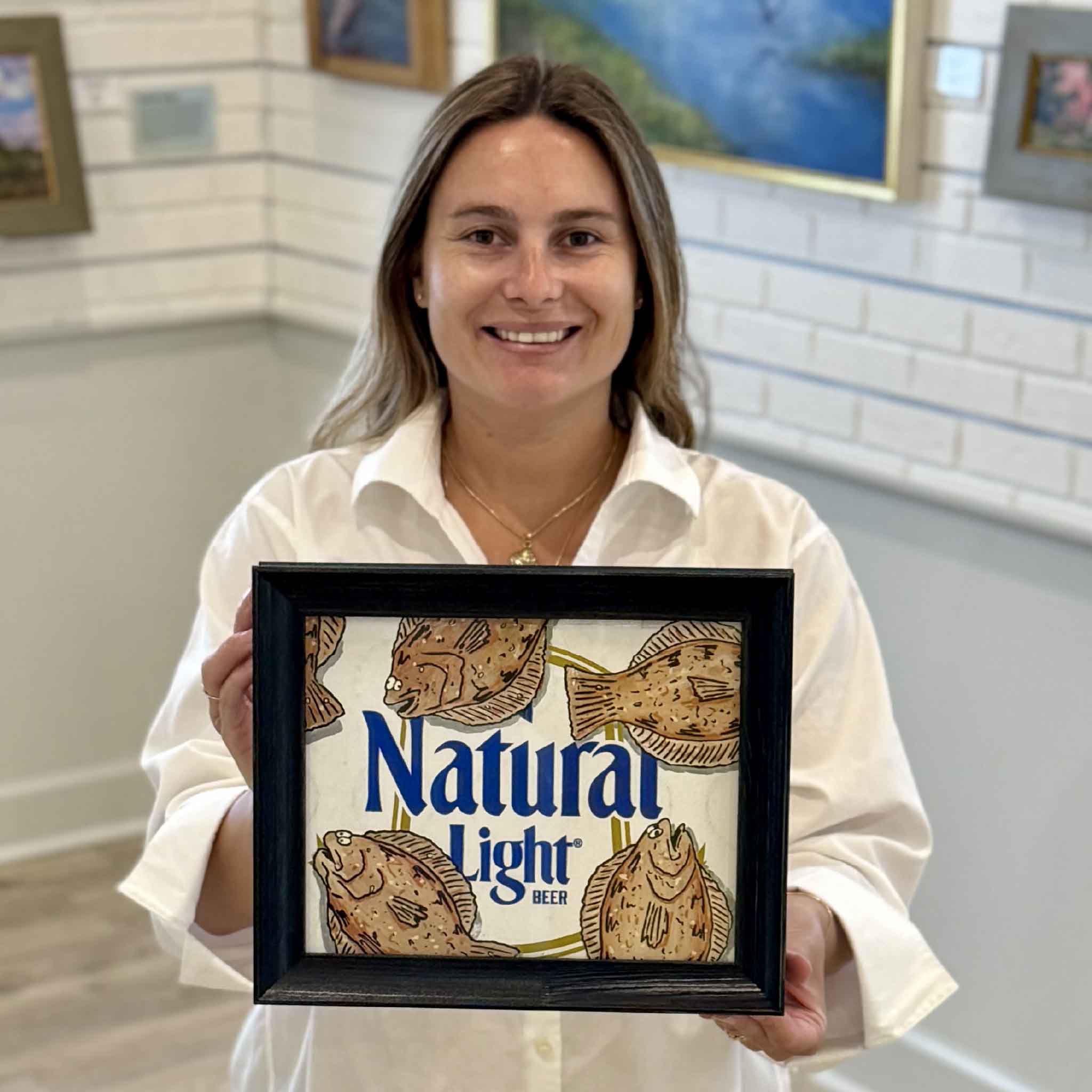 Kyra holds Floundahs 'n Poundahs, an original mixed media artwork by Charleston artist Nick Kawczynski featuring hand-painted flounder on a reclaimed Natural Light beer box. Professionally framed in navy blue and available at Gray Man Gallery in Pawleys Island, South Carolina.