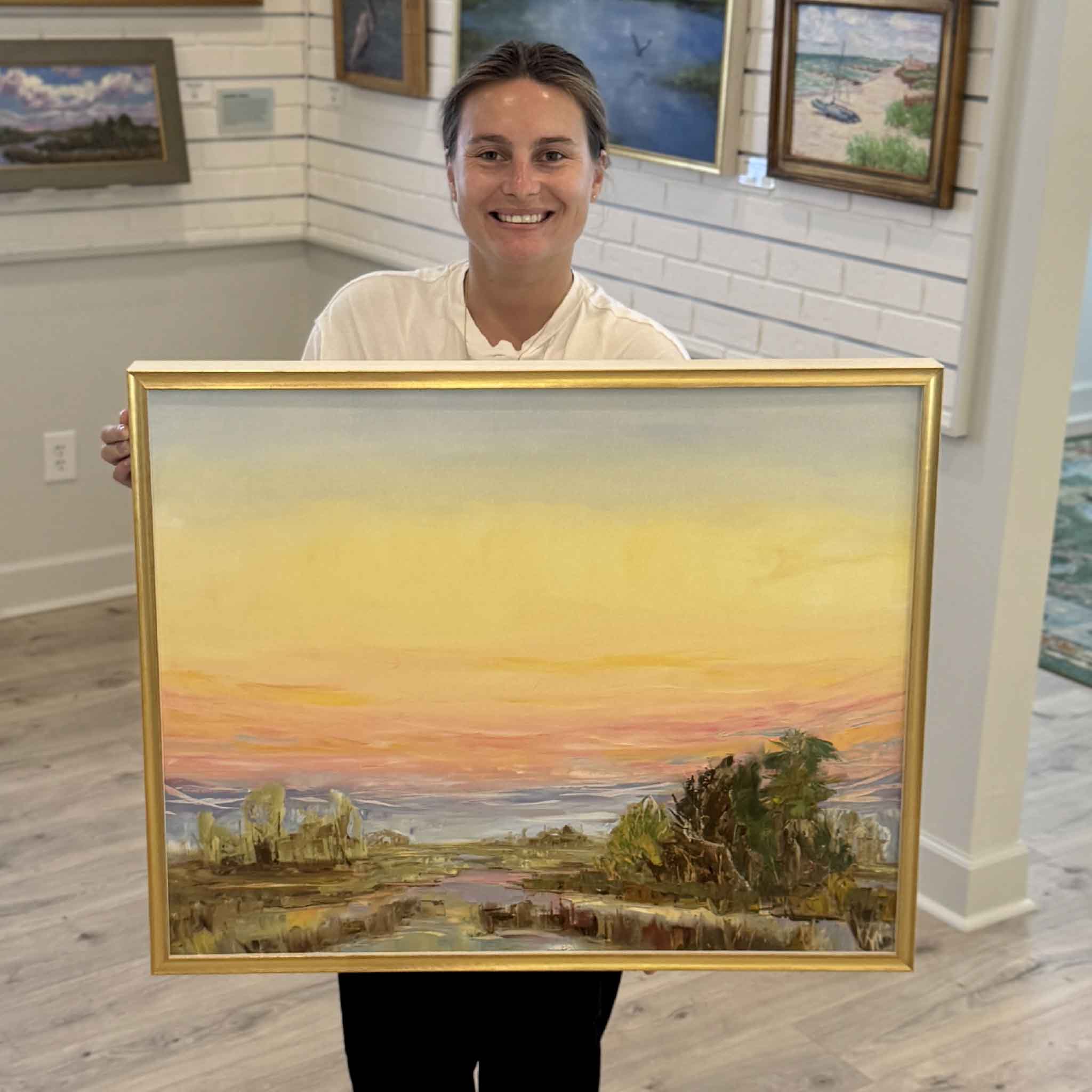 Gray Man Gallery team member Kyra holds Evening Grace, an original framed oil painting by Jeanne O'Neal featuring a luminous Lowcountry marsh at sunset with glowing pastel skies, reflective tidal waters, expressive brushwork, and Jeanne's signature gold frame, illustrating the artwork's scale for collectors and coastal art lovers.