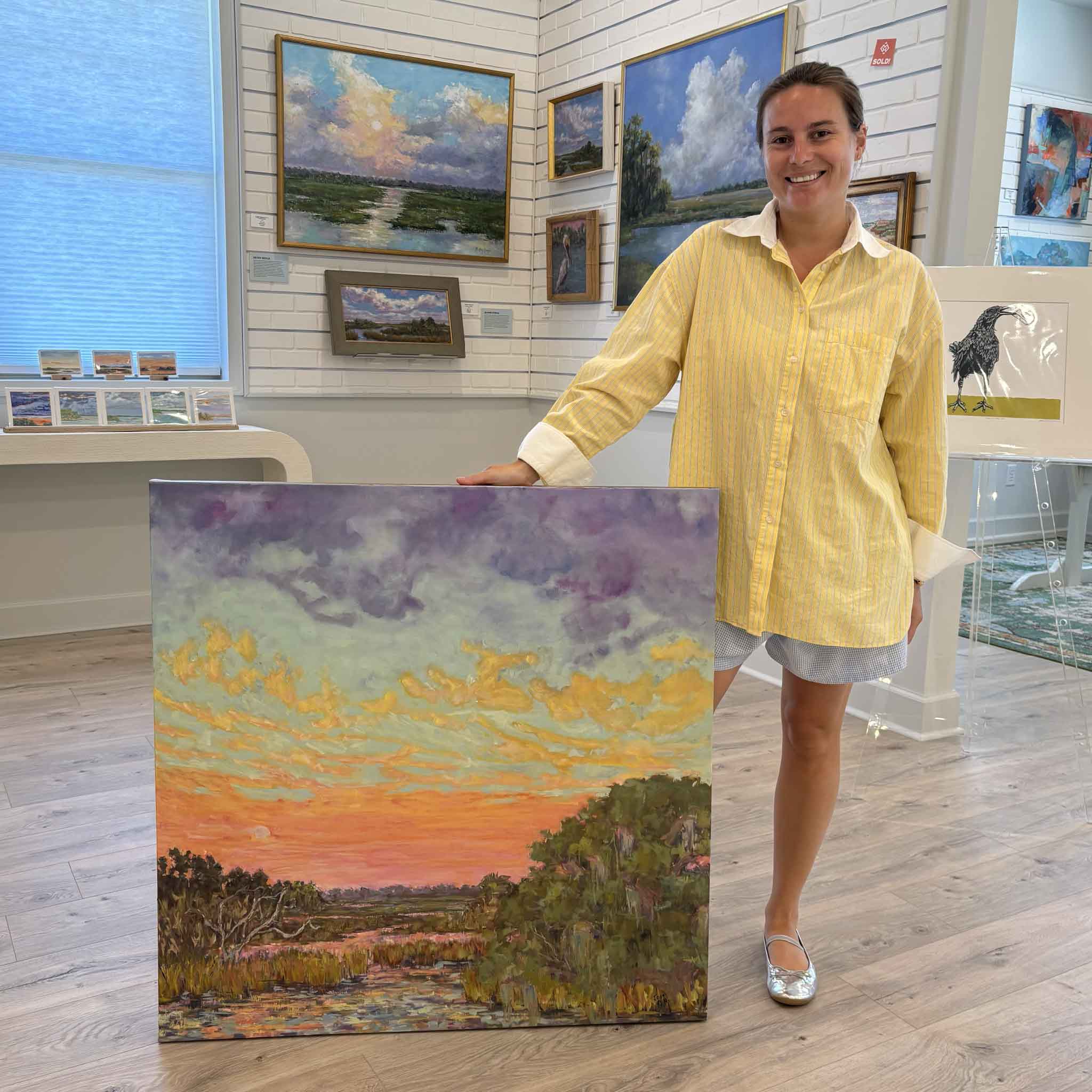 Kyra holds Hugs & Kisses: Afterglow, a 36x36 original oil painting by Jeanne O'Neal at Gray Man Gallery in Pawleys Island, South Carolina. The vibrant Lowcountry marsh landscape features an expansive sunset sky with luminous lavender, turquoise, coral, peach, and gold clouds above reflective tidal creeks, with gallery-wrapped edges finished in hand-painted gold.