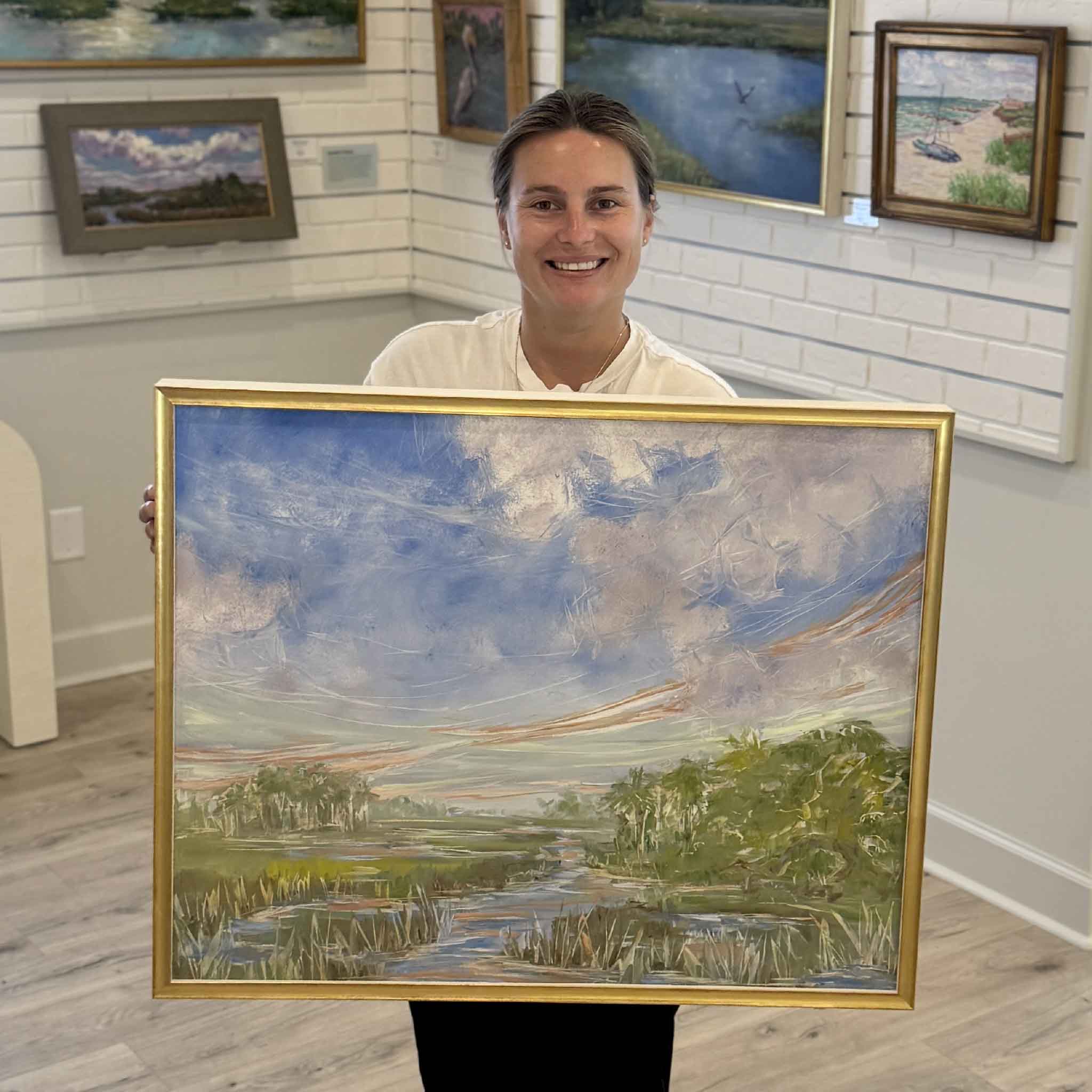 Gray Man Gallery team member Kyra holds Morning Promise, an original framed oil painting by Jeanne O'Neal. The expressive Lowcountry marsh landscape features sweeping blue skies, winding tidal creeks, lush marsh grasses, and Jeanne's signature gold frame, allowing customers to appreciate the artwork's impressive scale and vibrant painterly brushwork.