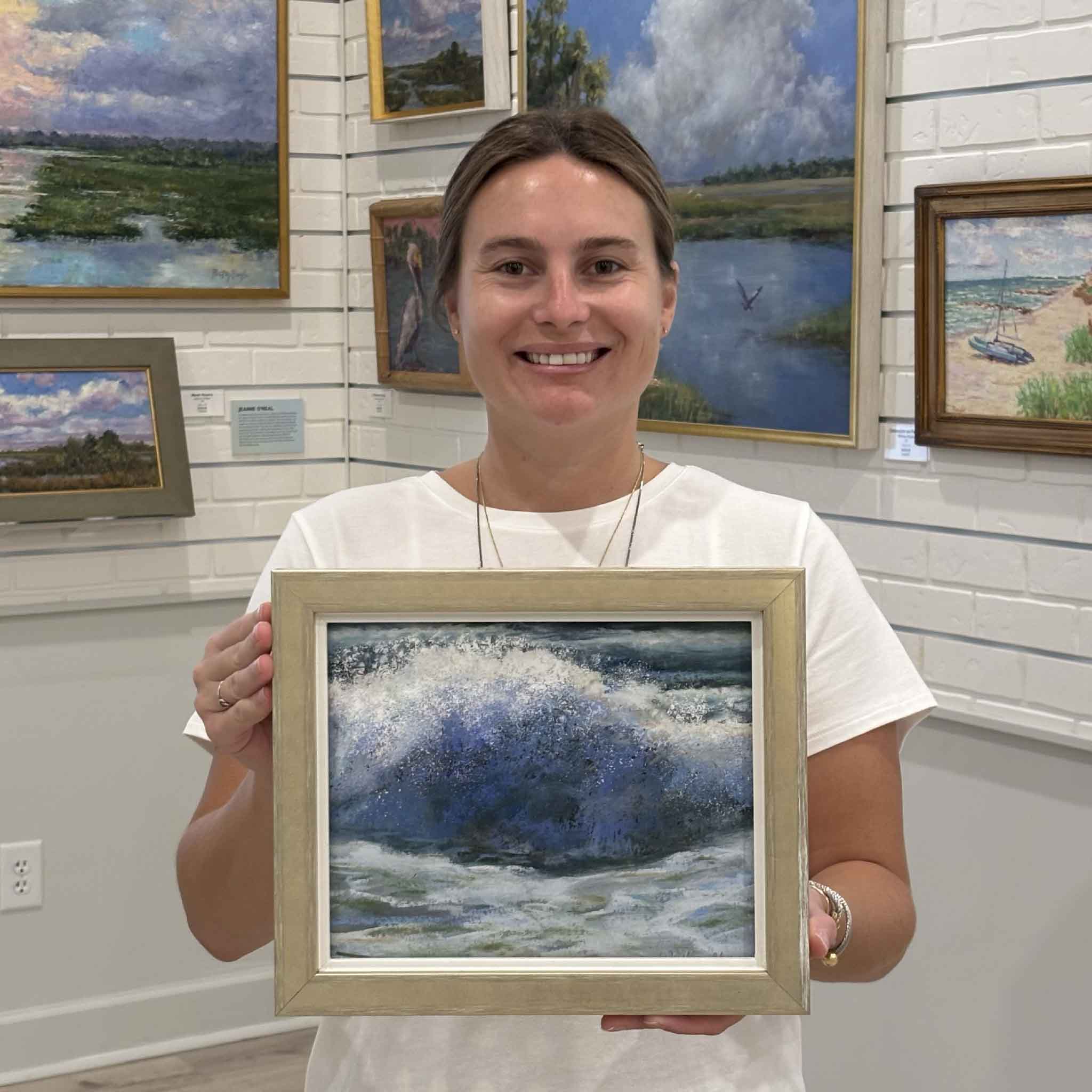 Kyra holds Exhilaration, an original soft pastel by local artist Joanne Willoughby featuring a dramatic Atlantic Ocean wave bursting with blue water and white sea spray at Gray Man Gallery in Pawleys Island, South Carolina.