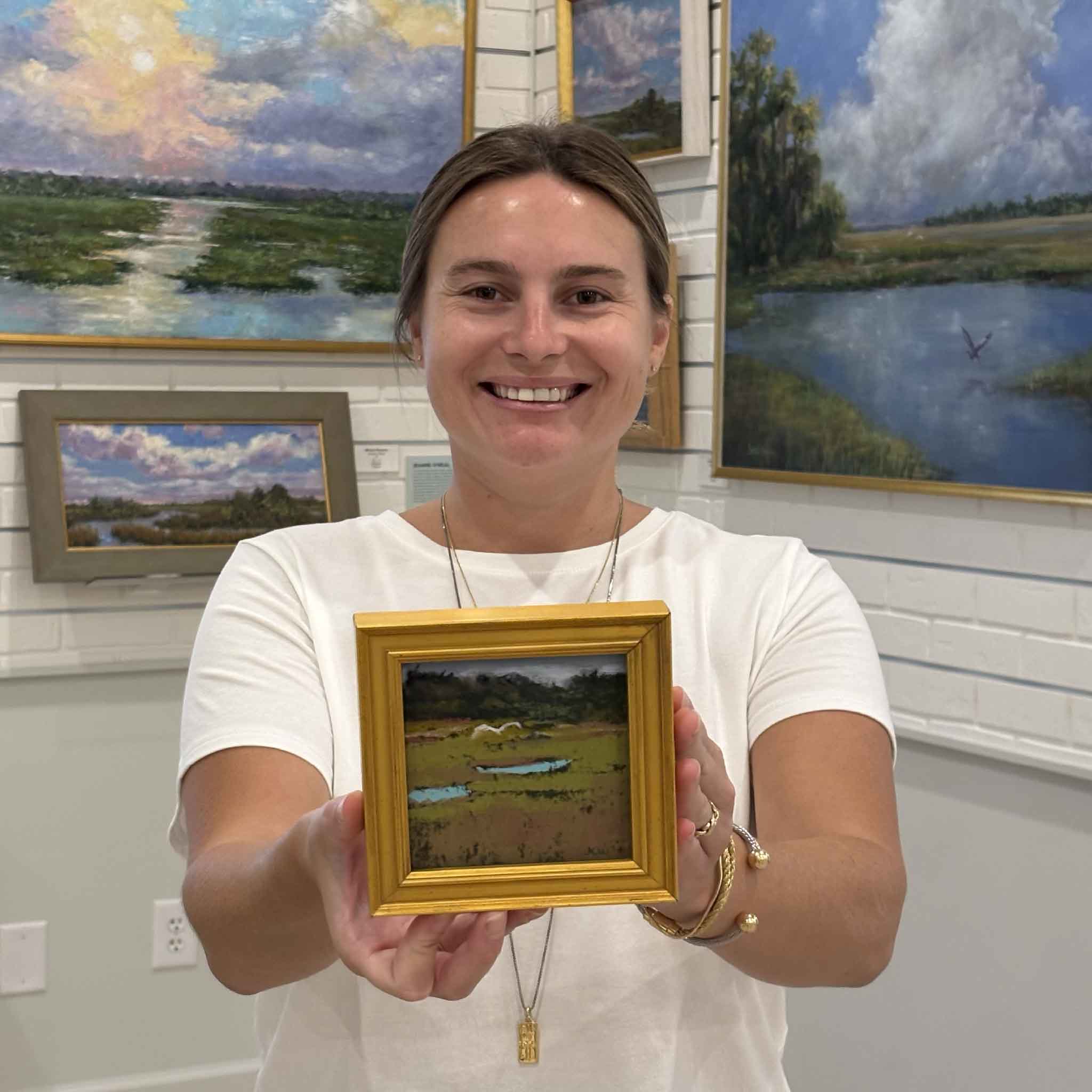 Kyra holds Over the Marsh, an original pastel painting by local artist Joanne Willoughby featuring two white egrets standing among reflective tidal pools in a tranquil South Carolina Lowcountry marsh. The artwork is professionally framed with Water White glass and available at Gray Man Gallery in Pawleys Island.