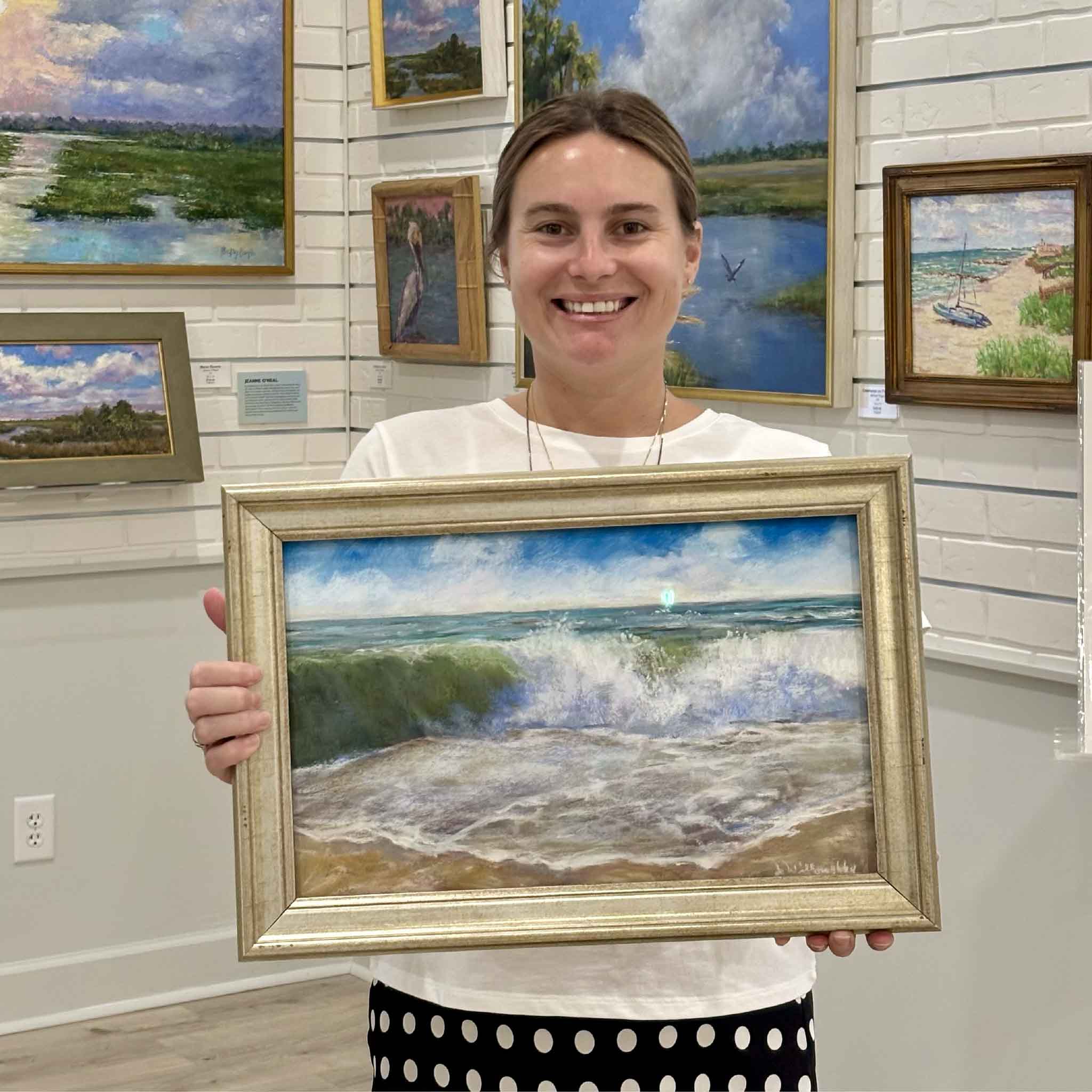 Kyra holds Perfect Day, an original ocean pastel painting by local artist Joanne Willoughby depicting a brilliant emerald wave breaking onto the shoreline beneath a blue coastal sky. Professionally framed with Water White glass, this original artwork is available at Gray Man Gallery in Pawleys Island, South Carolina.