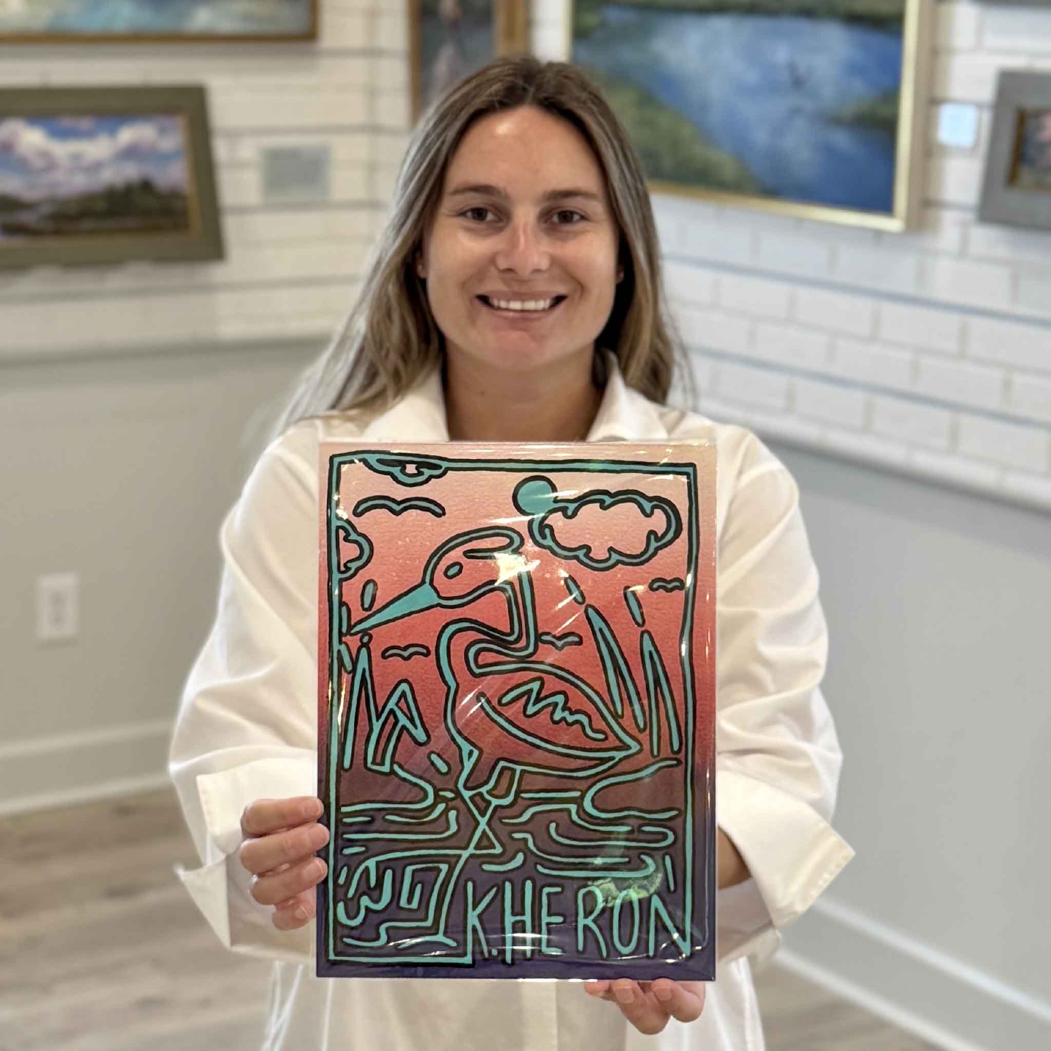 Kyra holds Keith Heron #1, an original mixed media artwork by Charleston artist Nick Kawczynski featuring a Great Heron rendered in a playful Keith Haring-inspired style. This colorful coastal artwork is available at Gray Man Gallery in Pawleys Island, South Carolina.