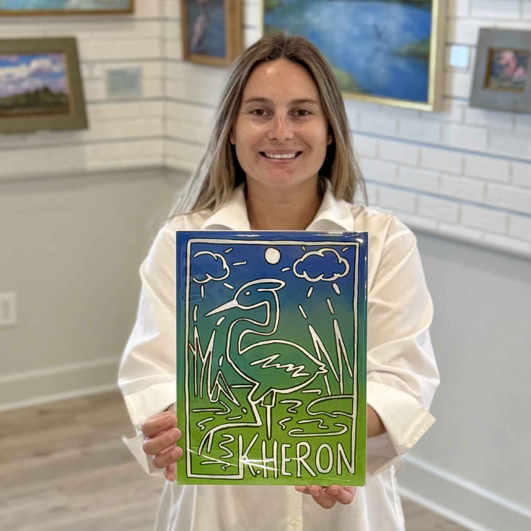 Kyra holds Keith Heron #2, an original mixed media artwork by Charleston artist Nick Kawczynski featuring a Great Heron rendered in a playful Keith Haring-inspired style with blue and green coastal colors. Available at Gray Man Gallery in Pawleys Island, South Carolina.