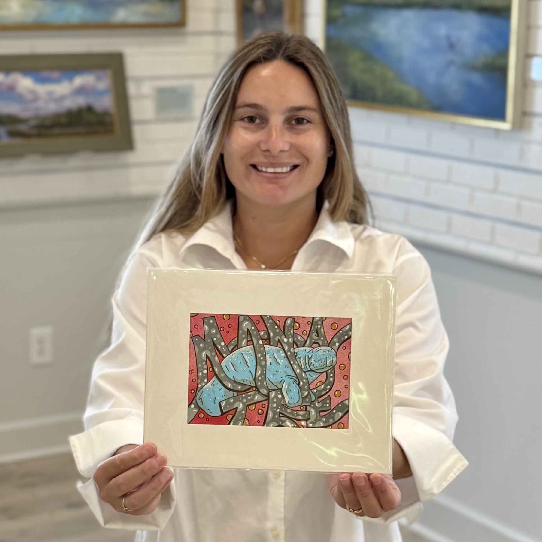 Kyra holds Manatee, an original mixed media artwork by Charleston artist Nick Kawczynski, at Gray Man Gallery in Pawleys Island, South Carolina. This colorful coastal piece features a bright blue manatee with hidden lettering woven throughout the composition and is professionally matted for display.