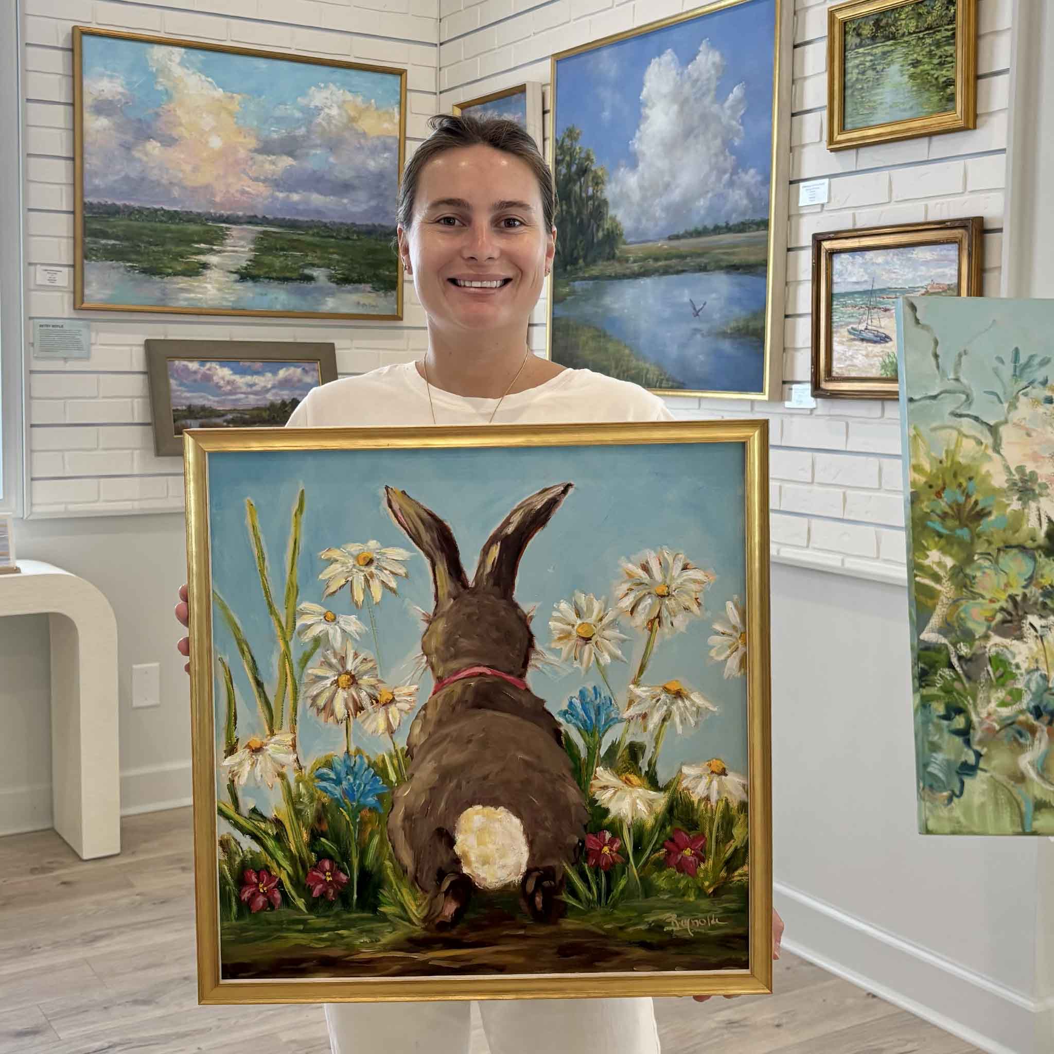 Kyra holds Out of the Rabbit Hole, an original oil painting by Jenny Reynolds, inside Gray Man Gallery in Pawleys Island, South Carolina. Professionally framed in an elegant gold frame and available at Gray Man Gallery in Pawleys Island, South Carolina, this whimsical original artwork brings joyful springtime charm to any home.