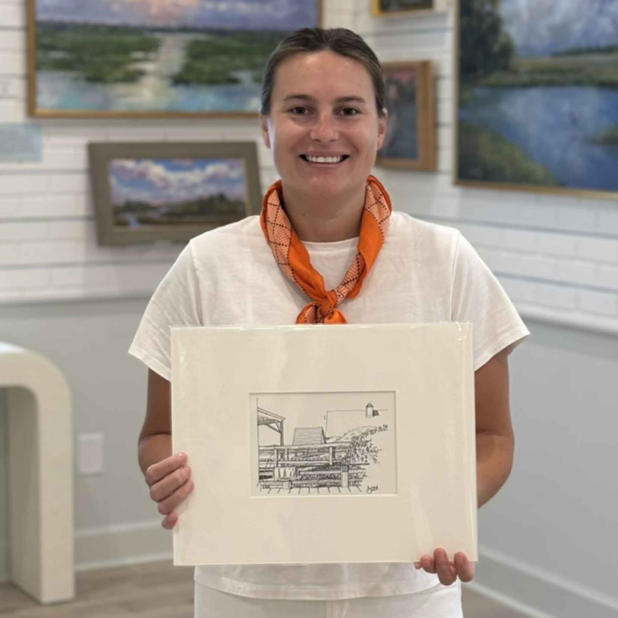 Kyra holding Rooftops Looking South by Todd Halamka at Gray Man Gallery in Pawleys Island, South Carolina. This professionally matted pen-and-ink print features a view of classic Pawleys Island beach cottages and fits a standard 11x14 frame for easy coastal decorating.
