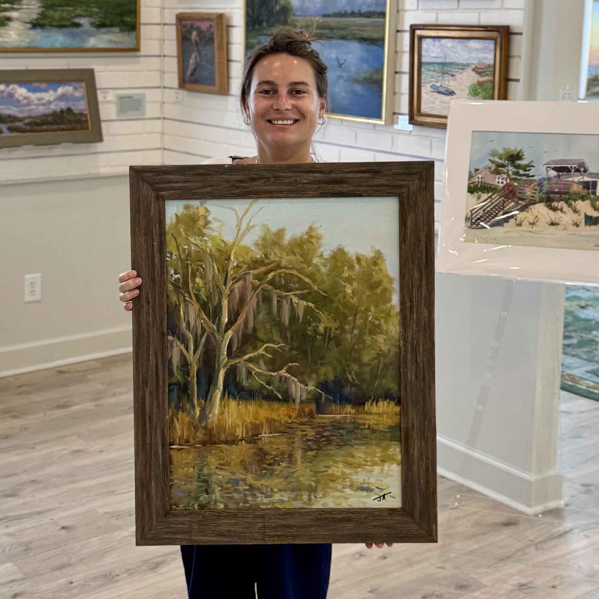 Kyra holds Still Waters, an original framed 18×24 Lowcountry oil painting by South Carolina local artist Jamie Arnold at Gray Man Gallery in Pawleys Island. The landscape features moss-draped live oaks, golden marsh grasses, and tranquil reflections that capture the peaceful beauty of the South Carolina coast.