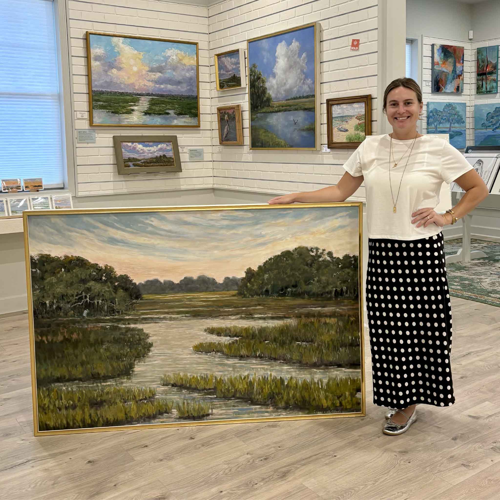 Kyra holds The Way Home, an original 60x40 Lowcountry marsh oil painting by Jeanne O'Neal, inside Gray Man Gallery in Pawleys Island, South Carolina. The photograph illustrates the impressive scale of this one-of-a-kind original, framed in Jeanne's signature handcrafted gold frame and featuring a winding tidal creek beneath an expansive coastal sky.
