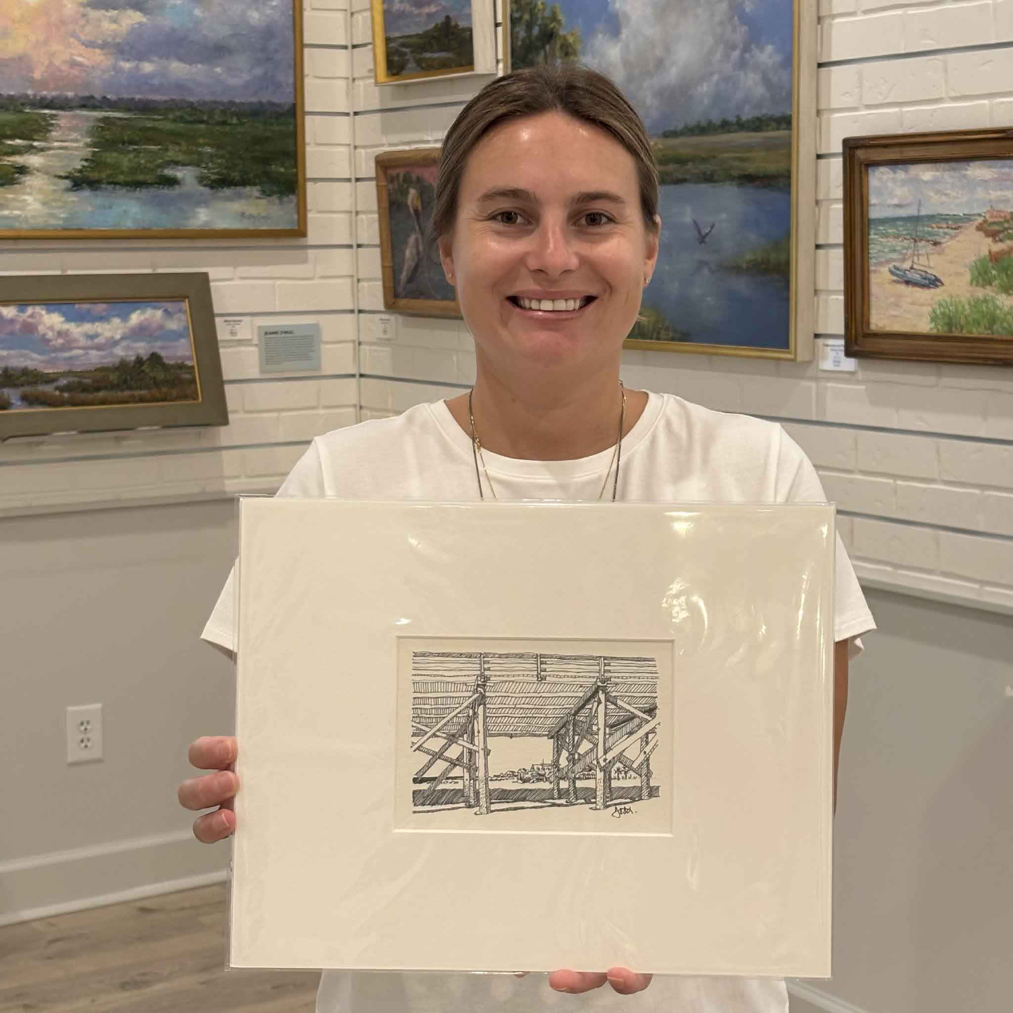 Kyra holds Todd Halamka's Beneath the Pawleys Pier print inside Gray Man Gallery in Pawleys Island, South Carolina. Professionally matted to a standard 11x14 size, this detailed pen-and-ink print celebrates the historic Pawleys Island Pier and is ready for one of our standard ready-made frames.