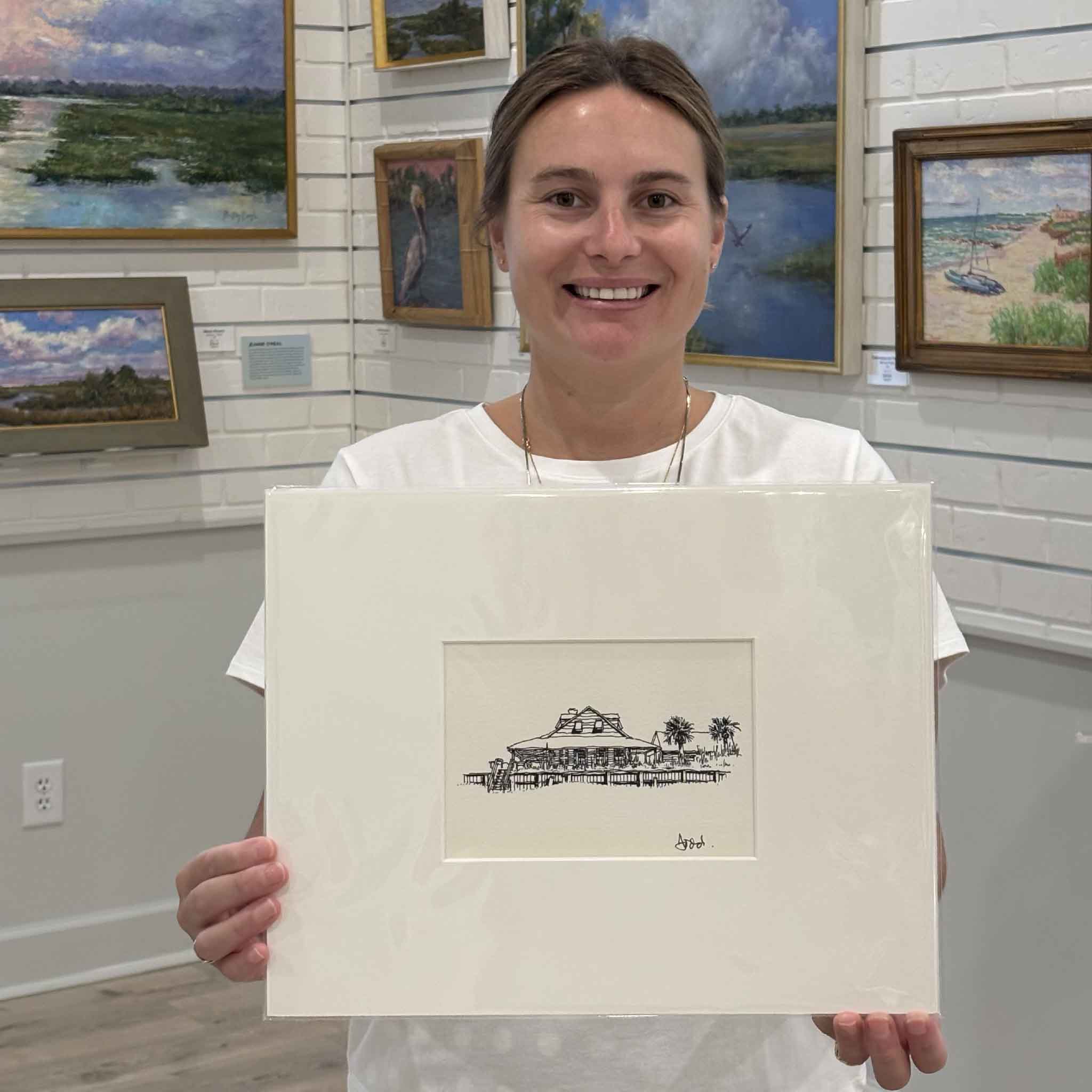 Kyra holds Todd Halamka's Caledonia at Pawleys Island pen-and-ink print at Gray Man Gallery in Pawleys Island, South Carolina. Professionally matted to a standard 11x14 size, this detailed print of the historic Caledonia Fish Club building is ready for one of our ready-made frames with no custom framing required.