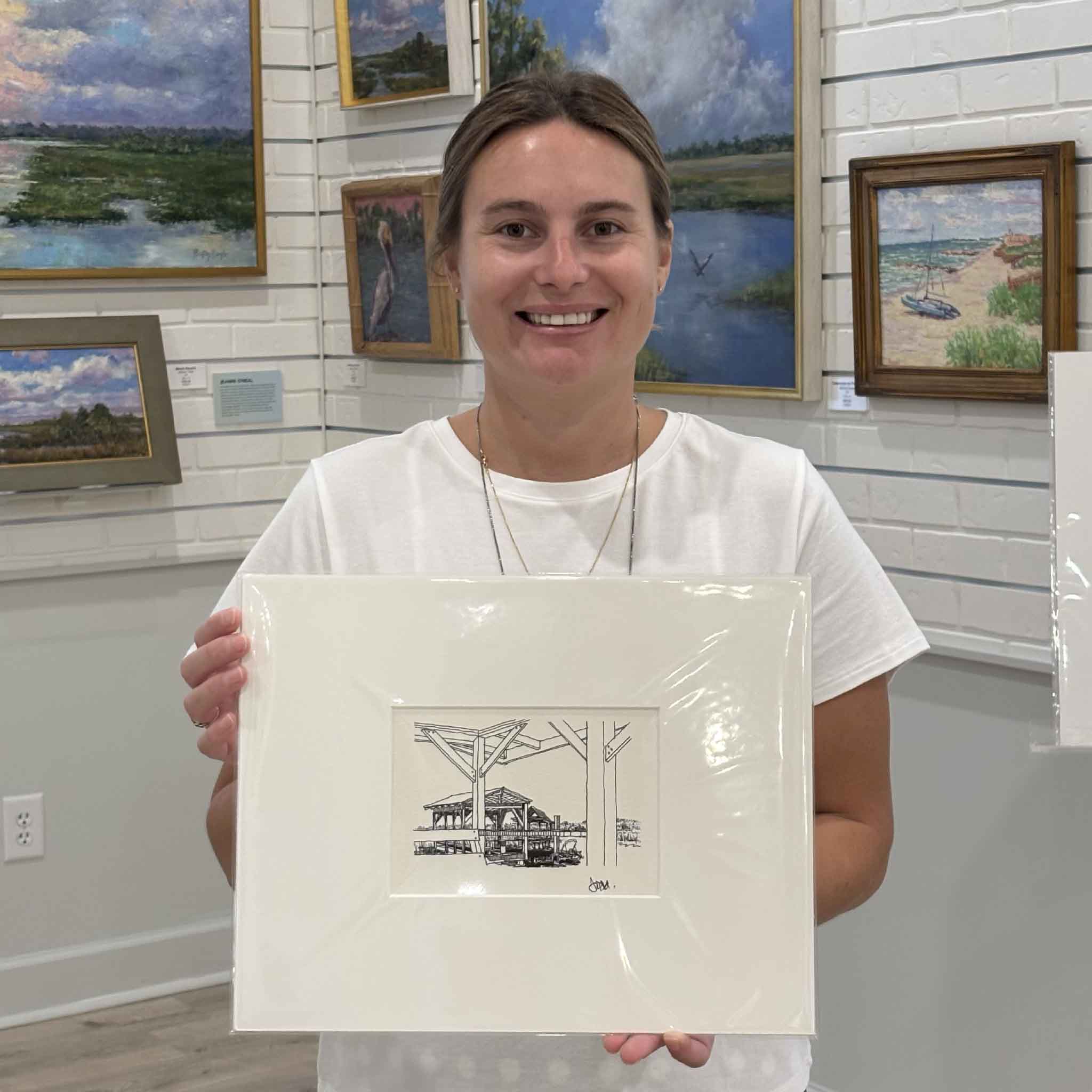 Kyra holds Todd Halamka's "Pawleys Island: Salt Marsh Looking South" pen-and-ink print at Gray Man Gallery in Pawleys Island, South Carolina. Professionally matted and available in standard 8x10 and 11x14 sizes, this detailed Lowcountry artwork captures a quiet marsh view through the framing of a dock, making it an easy-to-frame coastal gift or addition to any beach-inspired home.