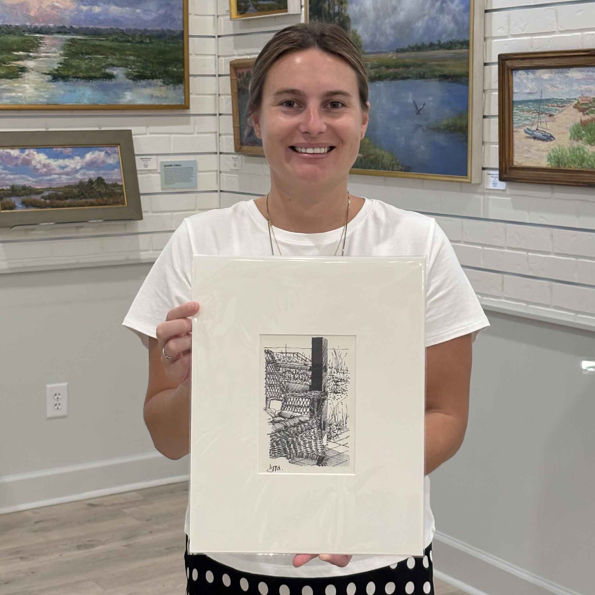 Kyra holds Pawleys Island Wicker Chair II, a professionally matted fine art print by Pawleys Island artist Todd Halamka, inside Gray Man Gallery in Pawleys Island, South Carolina. The photo shows the scale of the detailed pen-and-ink print depicting an iconic wicker porch chair overlooking the dunes, ready to fit a standard ready-made frame without custom framing.