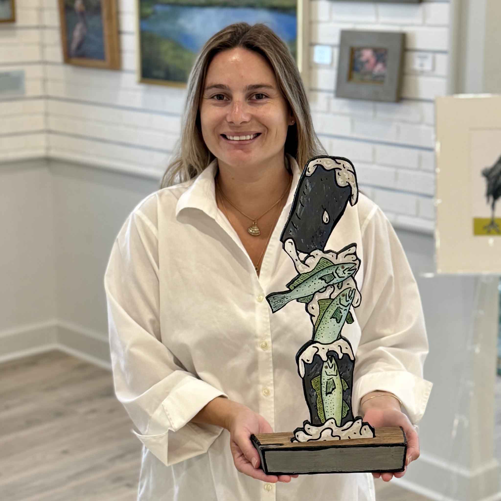 Kyra holds Trouts 'n Stouts, an original mixed media sculpture by Charleston artist Nick Kawczynski made from reclaimed marine-grade plywood and found wood, at Gray Man Gallery in Pawleys Island, South Carolina.