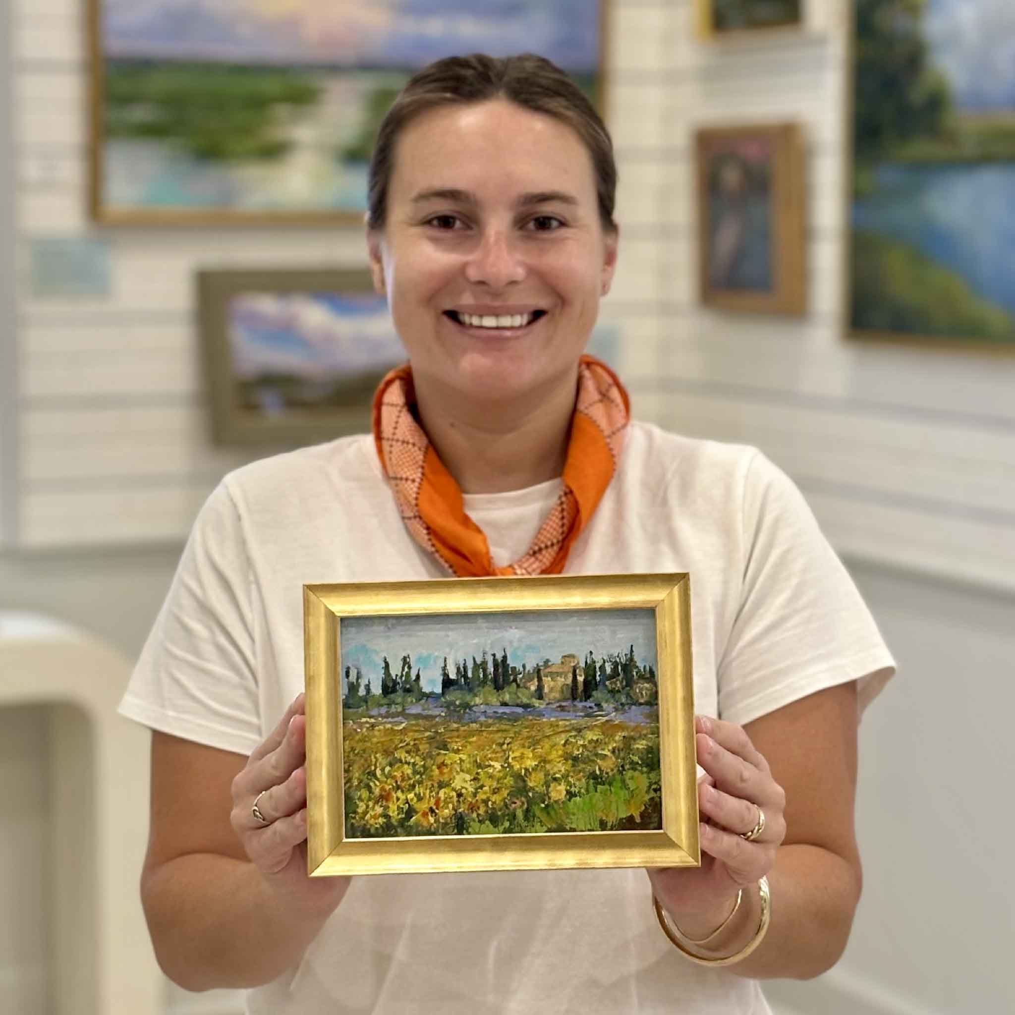 Gray Man Gallery team member Kyra holding Tuscan Spring by Jeanne O'Neal to show scale, an original oil painting of a colorful Tuscan countryside landscape framed in Jeanne's signature gold frame.
