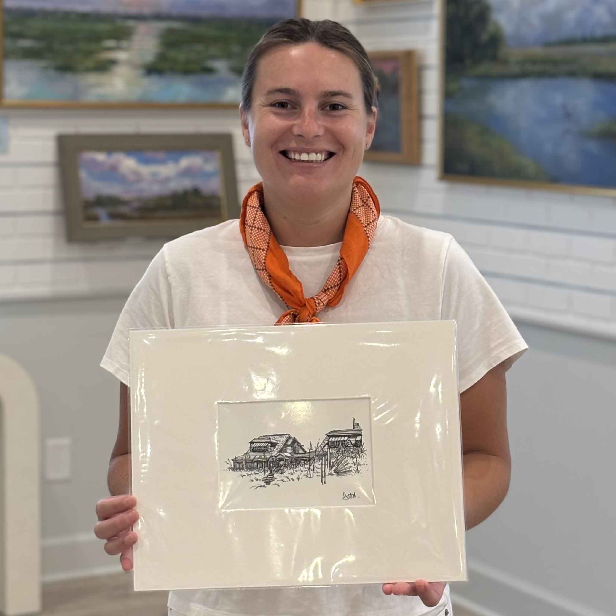 Kyra holding Two Beach Houses III by Todd Halamka at Gray Man Gallery in Pawleys Island, South Carolina. This professionally matted pen-and-ink print depicts traditional Pawleys Island beach cottages and fits a standard 11x14 frame, making it an easy gift for coastal homeowners and Lowcountry art collectors.