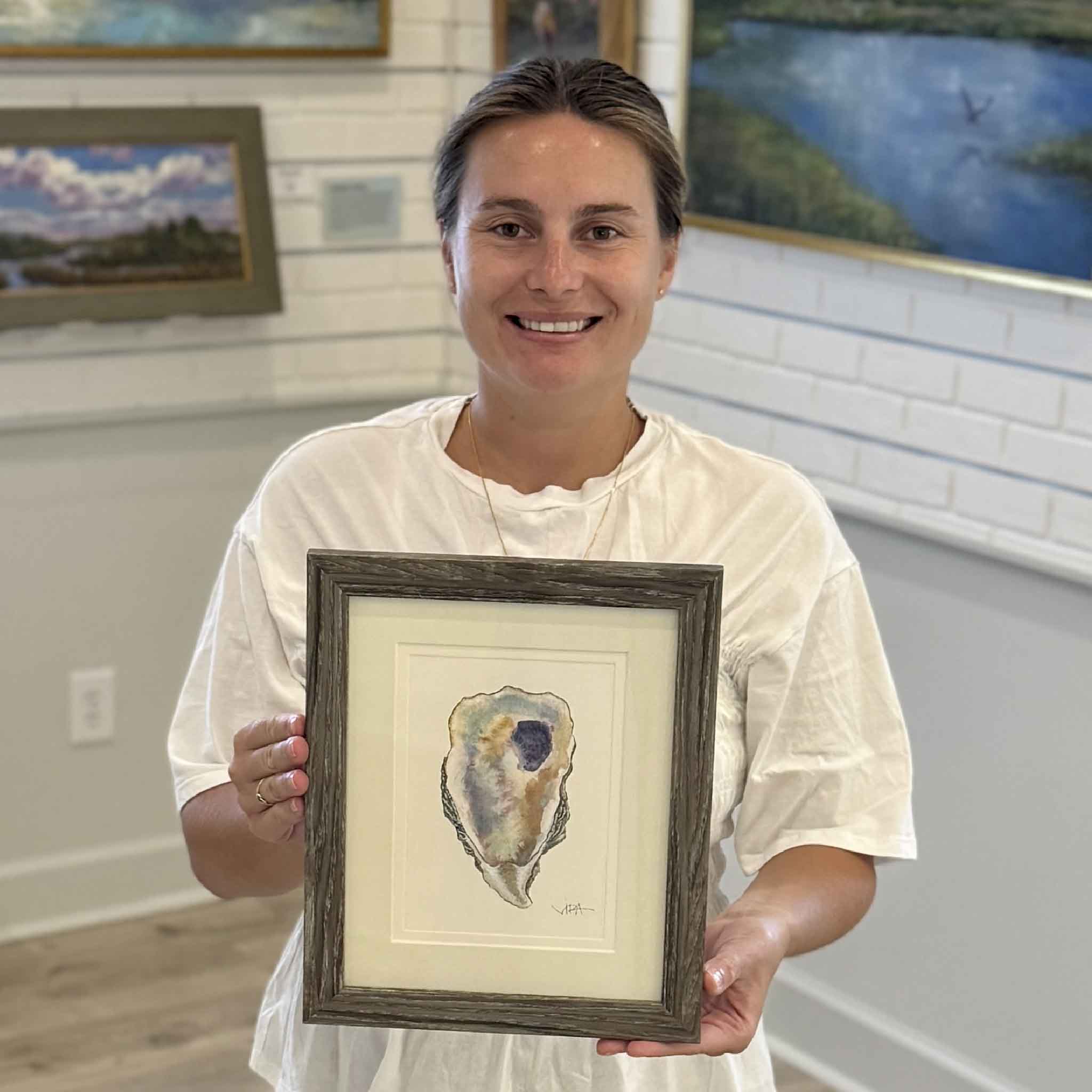 Kyra holds Pawleys Oyster Shell VI, an original watercolor by local Pawleys Island artist Vida Miller, professionally double matted and framed in a weathered gray wood frame. The one-of-a-kind oyster shell painting features soft coastal blues, pearl whites, warm golden hues, and a rich lavender interior, available at Gray Man Gallery in Pawleys Island, South Carolina.