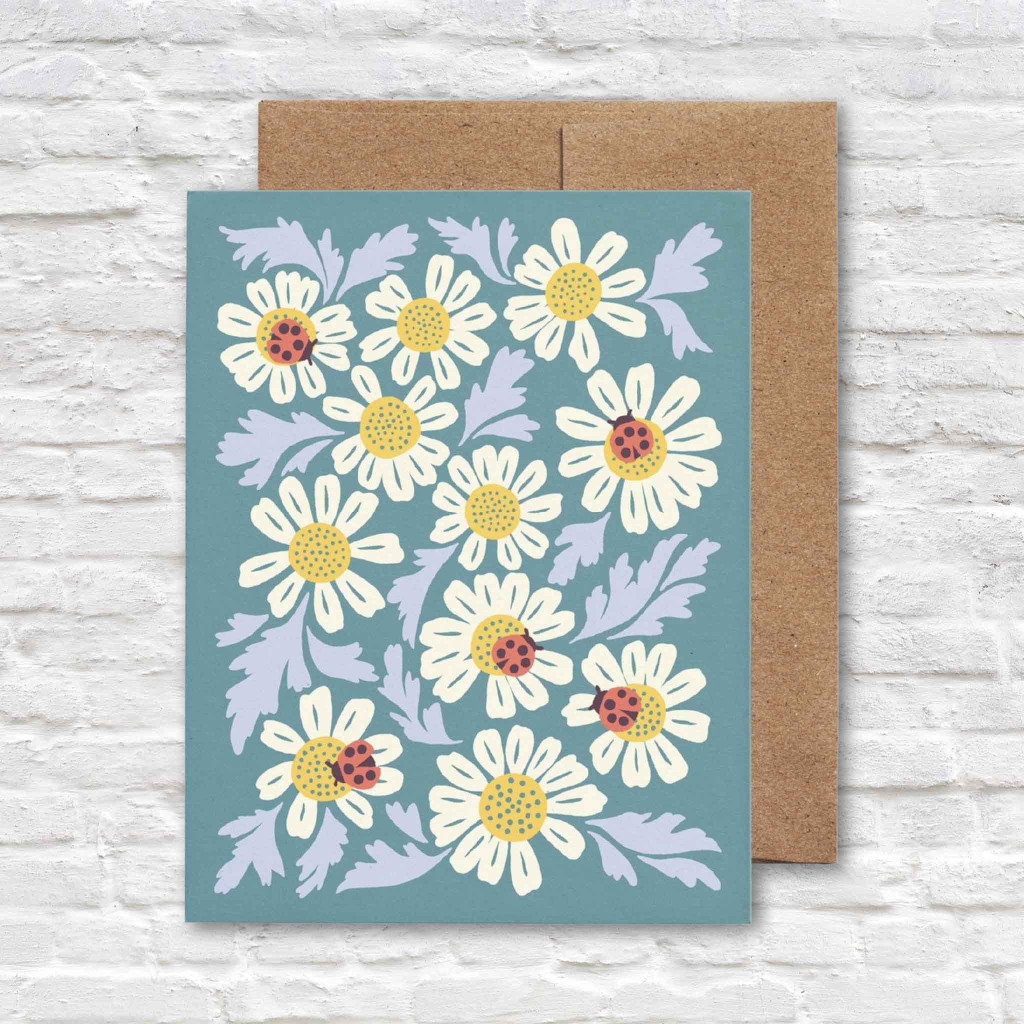 Ladybugs & Chamomile greeting card by Chrissie Van Hoever featuring white chamomile blossoms with bright red ladybugs on a soft blue background, paired with a recycled kraft envelope. Available at Gray Man Gallery in Pawleys Island, South Carolina.