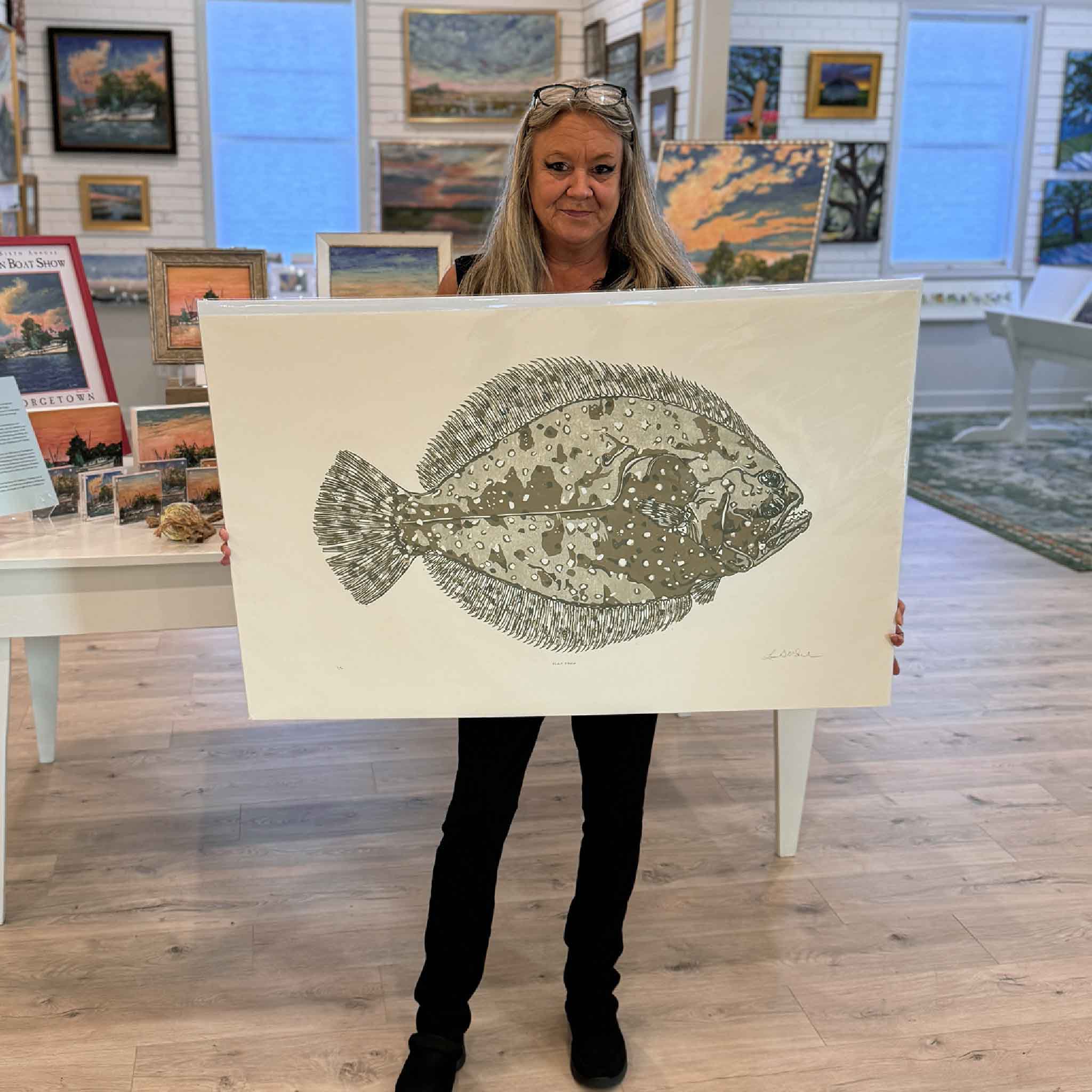 Charlotte holding “Flat Fred,” an original linocut by Laurie McIntosh featuring a detailed flounder green tones at Gray Man Gallery.