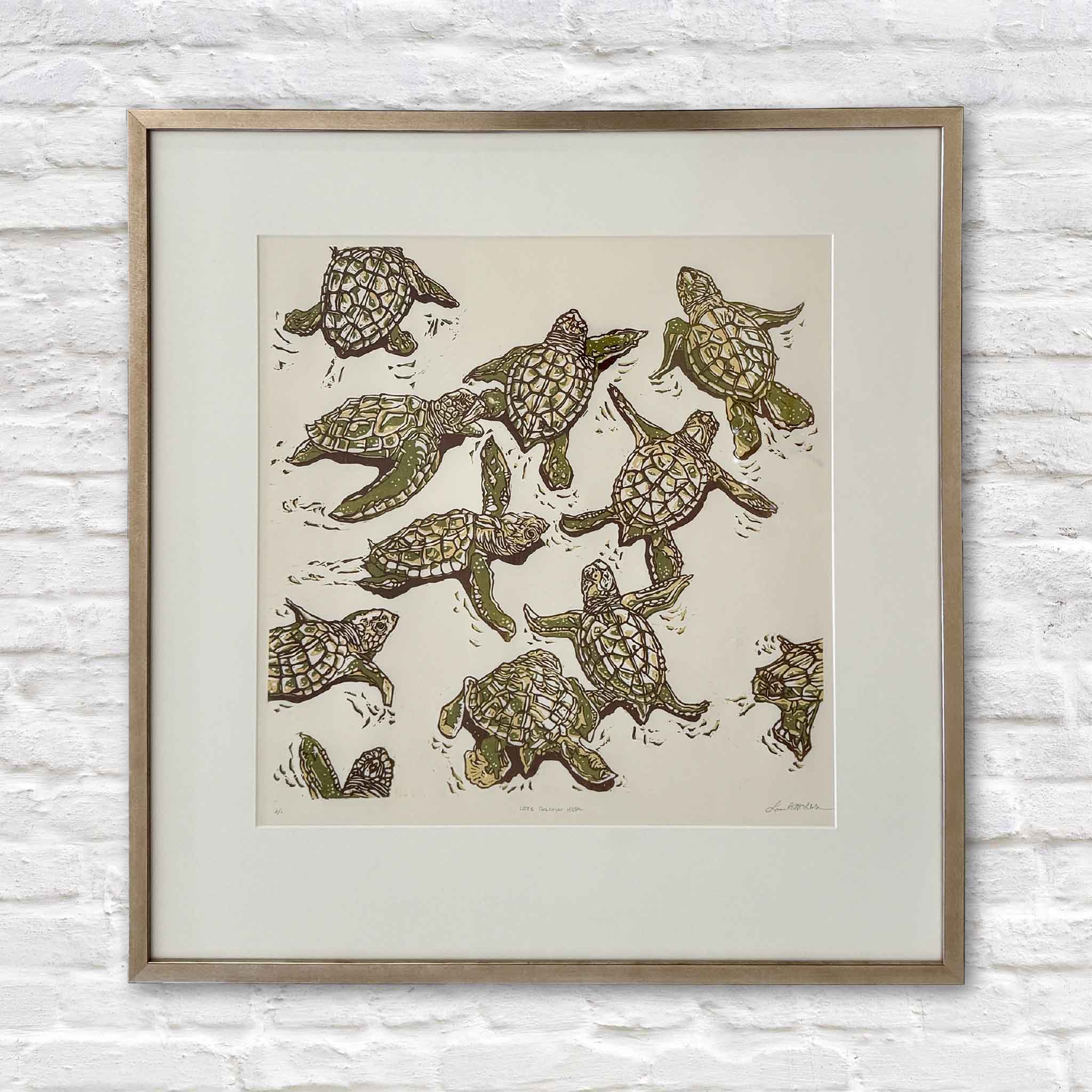 Let’s Follow Her original reduction linocut by Lowcountry artist Laurie McIntosh, featuring a group of young sea turtles hand-printed in earthy greens and browns, professionally matted and finished in the artist’s signature champagne frame at Gray Man Gallery in Pawleys Island, South Carolina.