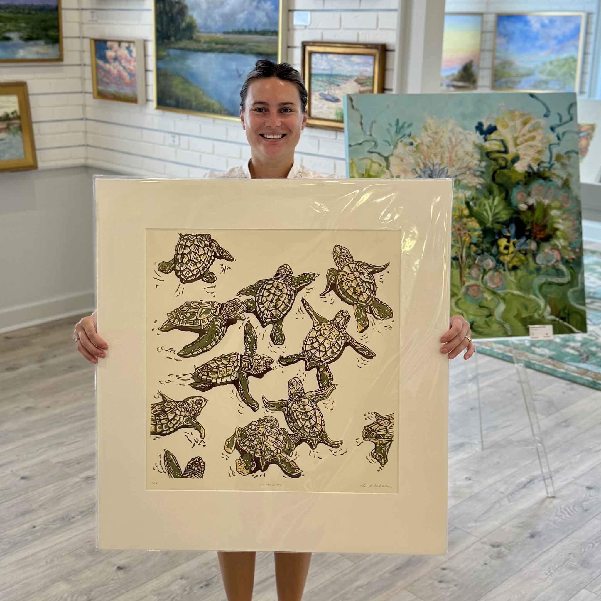 Kyra holding Let’s Follow Her, an original hand-pulled reduction linocut by Laurie McIntosh featuring a lively group of sea turtles in layered olive green, ochre and brown, shown matted and ready for framing inside Gray Man Gallery in Pawleys Island, South Carolina.