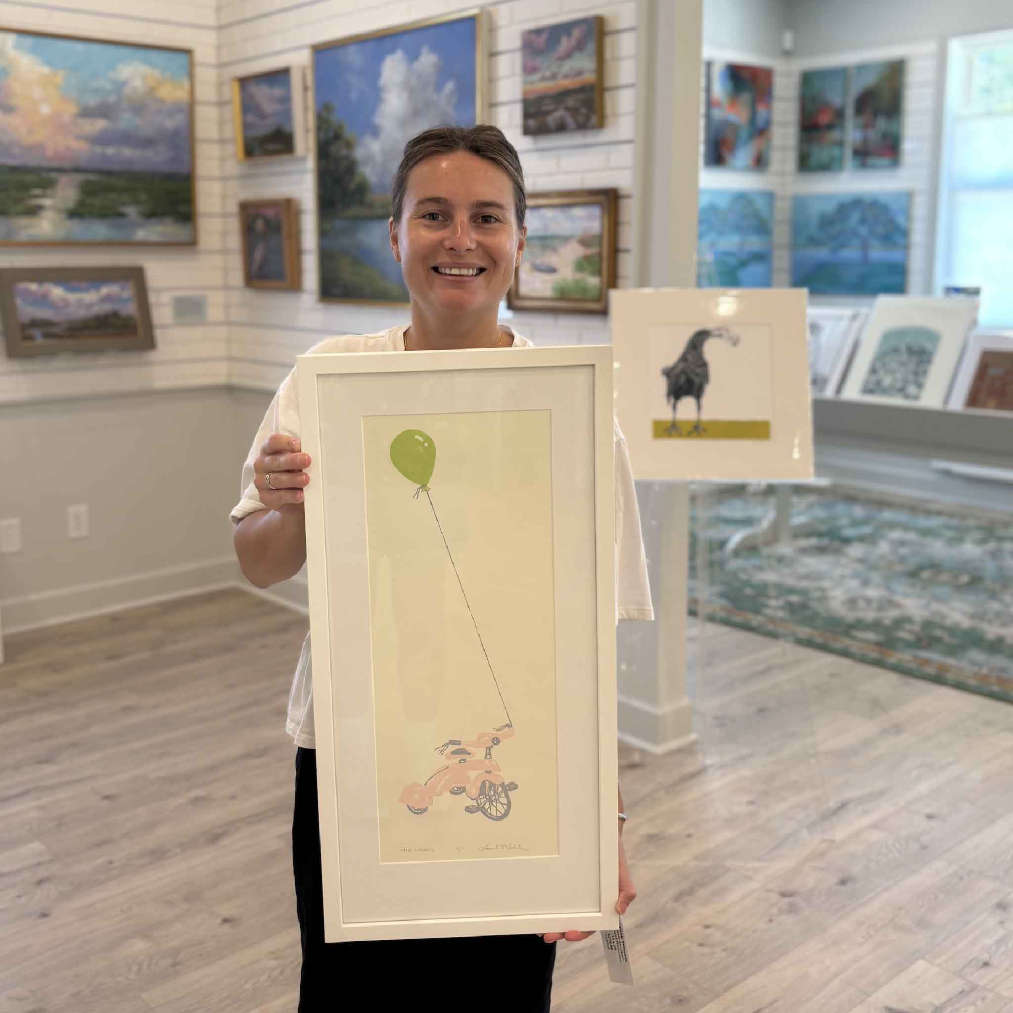 Kyra holds Pink Wheels, an original hand-pulled reduction linocut by South Carolina artist Laurie McIntosh, at Gray Man Gallery in Pawleys Island. This whimsical original print features a vintage pink tricycle tethered to a bright green balloon floating overhead, showcasing the artwork's scale, custom white frame, and playful color palette. Available at Gray Man Gallery, this one-of-a-kind original linocut is created using Laurie McIntosh's signature reduction process, making every impression unique.
