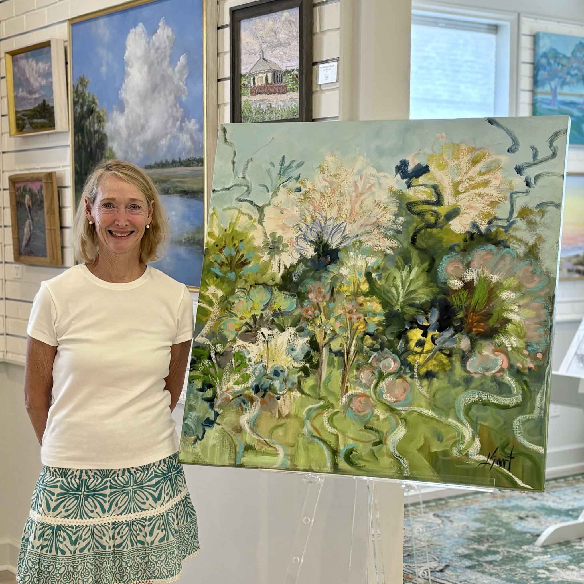 Artist Libby Smart stands beside her original oil painting Glorious Sea Fans at Gray Man Gallery in Pawleys Island, South Carolina. This vibrant botanical-inspired contemporary floral painting features soft greens, blues, creams, and blush tones with expressive brushwork and flowing organic forms inspired by Lowcountry sea fans and coastal gardens. Available exclusively at Gray Man Gallery, the image showcases the artist with her original artwork in the gallery.