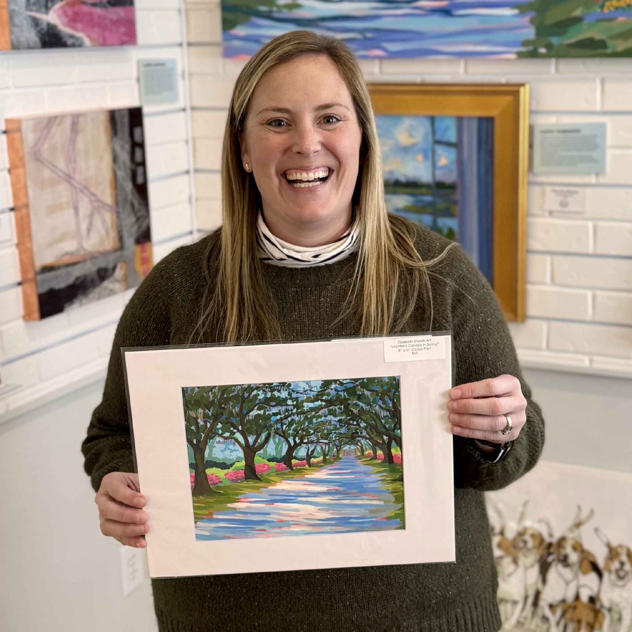 Elizabeth Sheats Litchfield Canopy in Spring fine art giclée print matted to fit 11x14 frame, held by staff inside Gray Man Gallery in Pawleys Island South Carolina, featuring live oak canopy at Litchfield Plantation with Spanish moss, pink spring azaleas, sunlit oak-lined drive, classic Lowcountry landscape, coastal Southern wall art, and locally curated artwork