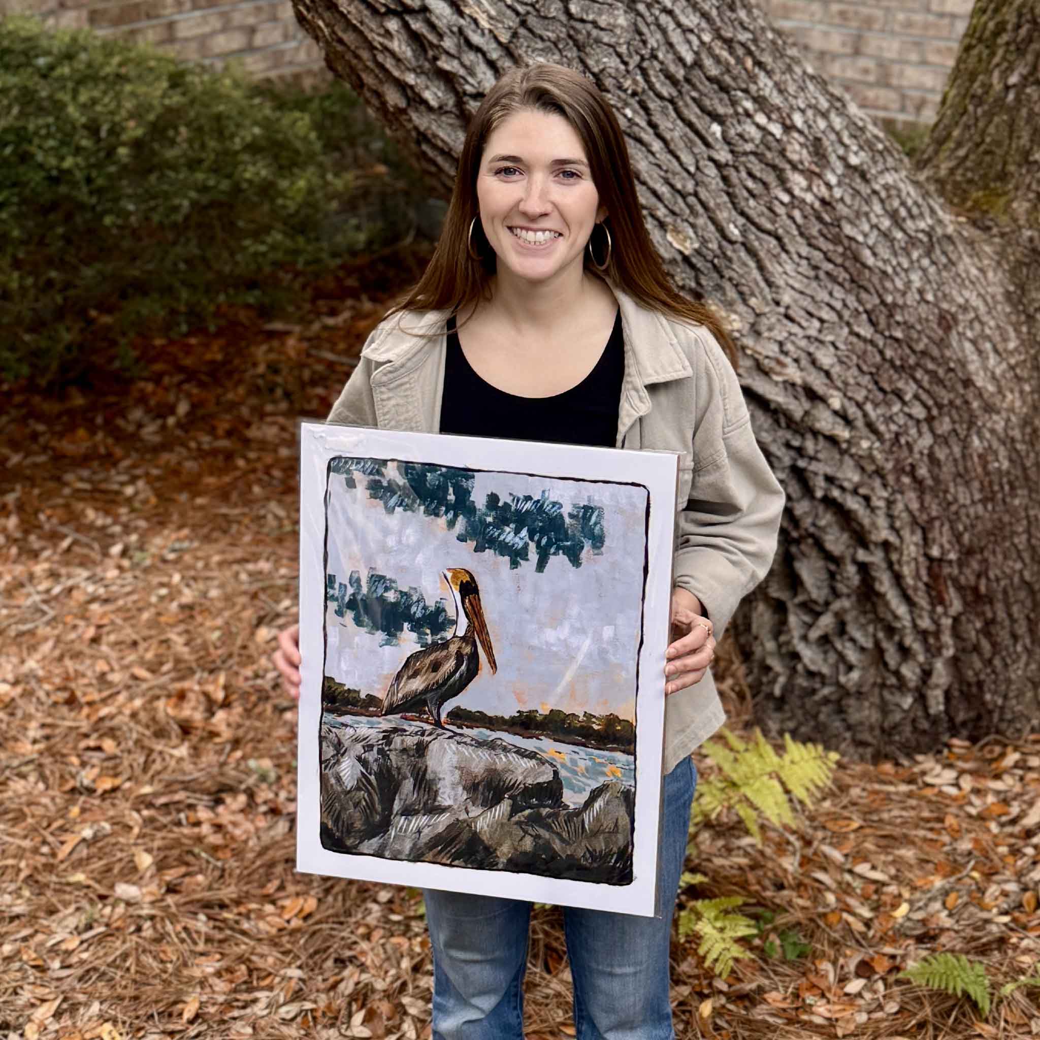 Maggie Pelton, local artist, holding her Brown Pelican fine art print outdoors, featuring a pelican perched along the South Carolina coast with expressive brushwork and muted coastal tones, photographed near Pawleys Island and available at Gray Man Gallery in Pawleys Island, South Carolina