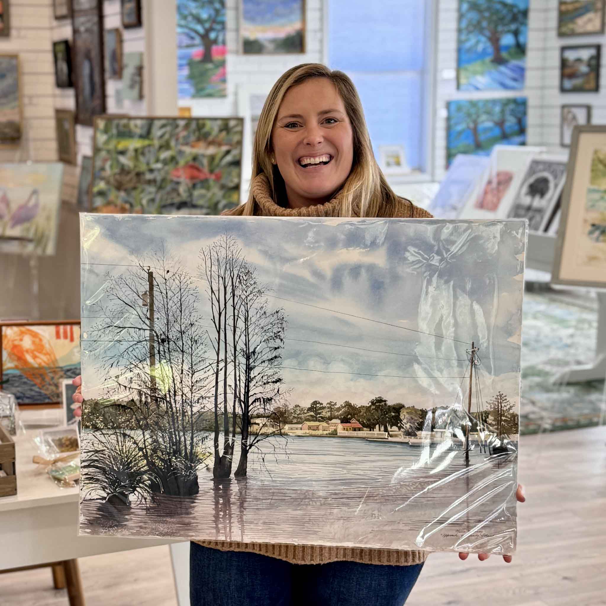 Maura Kenny, local artist, holding her original watercolor Approach to Sandy Island inside Gray Man Gallery, featuring a tranquil waterway view inspired by Sandy Island near Pawleys Island, South Carolina