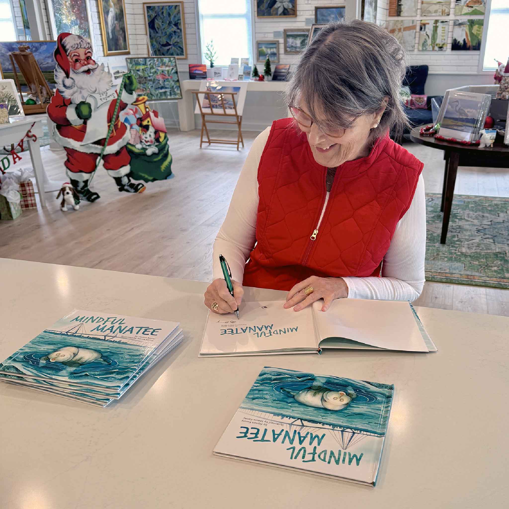 Illustrator Maura Kenny sitting at a table in Gray Man Gallery signing copies of Mindful Manatee during a book signing event.
