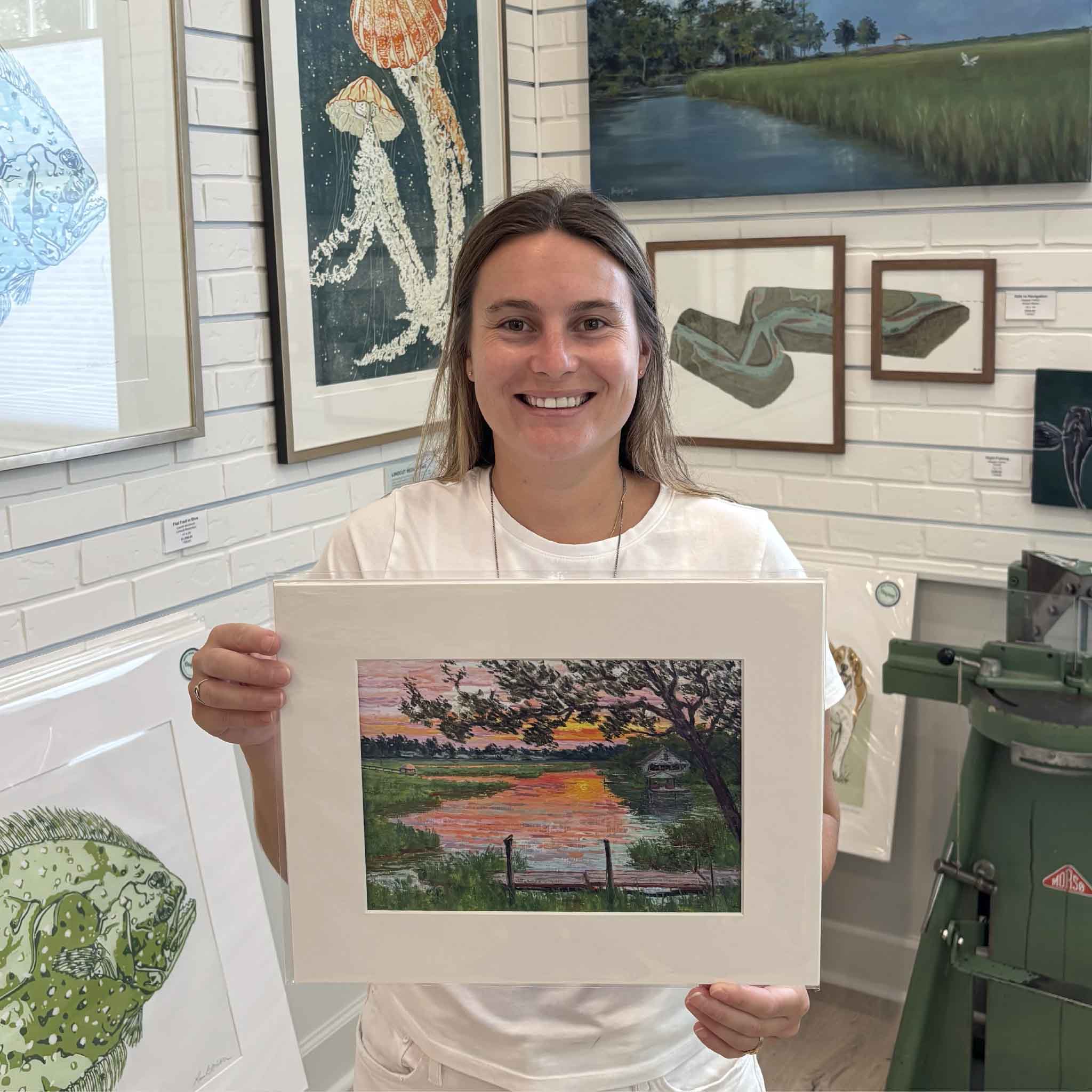Kyra holds the 11×14 matted Fading Light giclée print by Michael Rogan at Gray Man Gallery in Pawleys Island, South Carolina, featuring Rogan’s Lowcountry marsh sunset painted in South Litchfield.