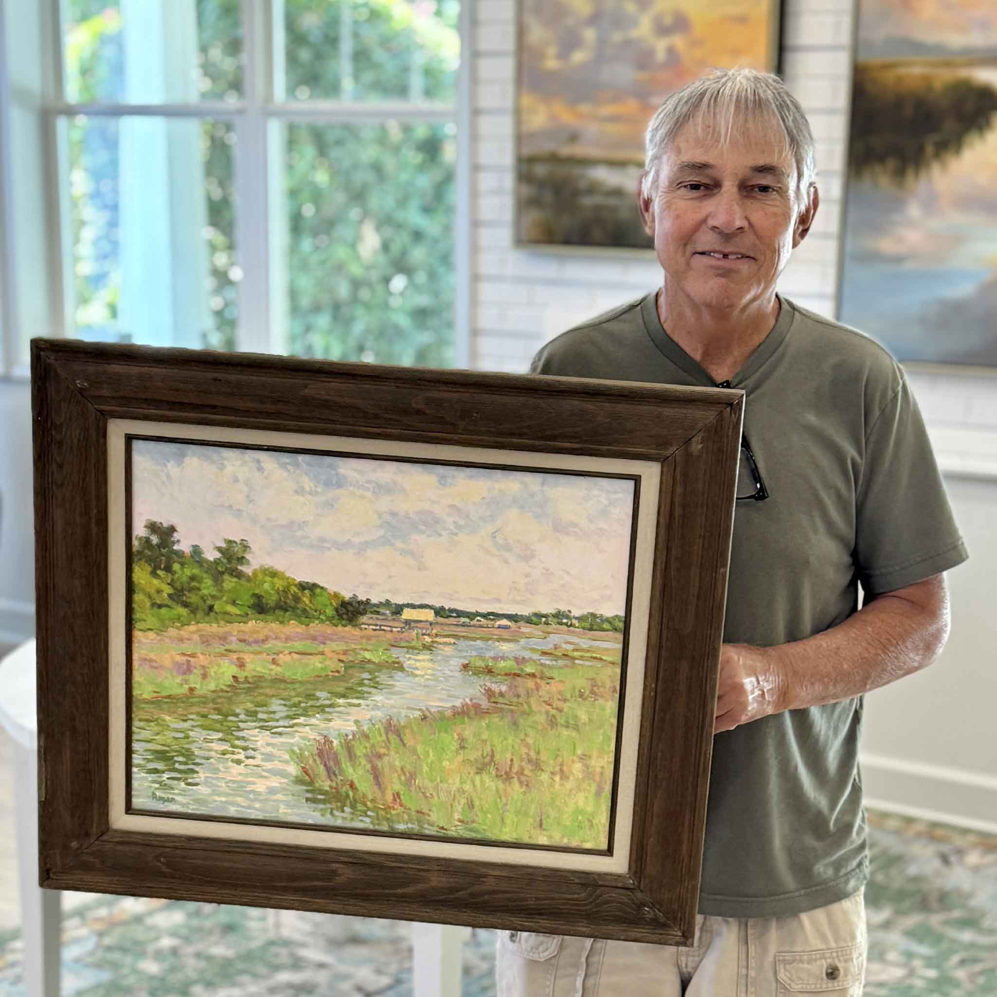 Local artist Michael Rogan holding Spring at Litchfield, a framed original oil painting inspired by the marsh landscapes of Litchfield, South Carolina, featuring fresh spring greens, reflective tidal water, and soft atmospheric Lowcountry light at Gray Man Gallery