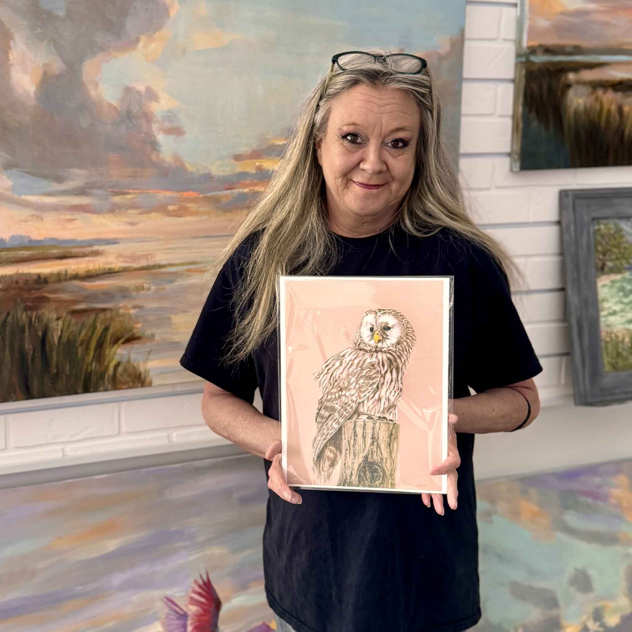 Charlotte holding Mimi Beaver’s owl watercolor print showing detailed feathers and warm peach background, used as the cover image for the Mimi Beaver Giving Collection benefiting senior dogs.