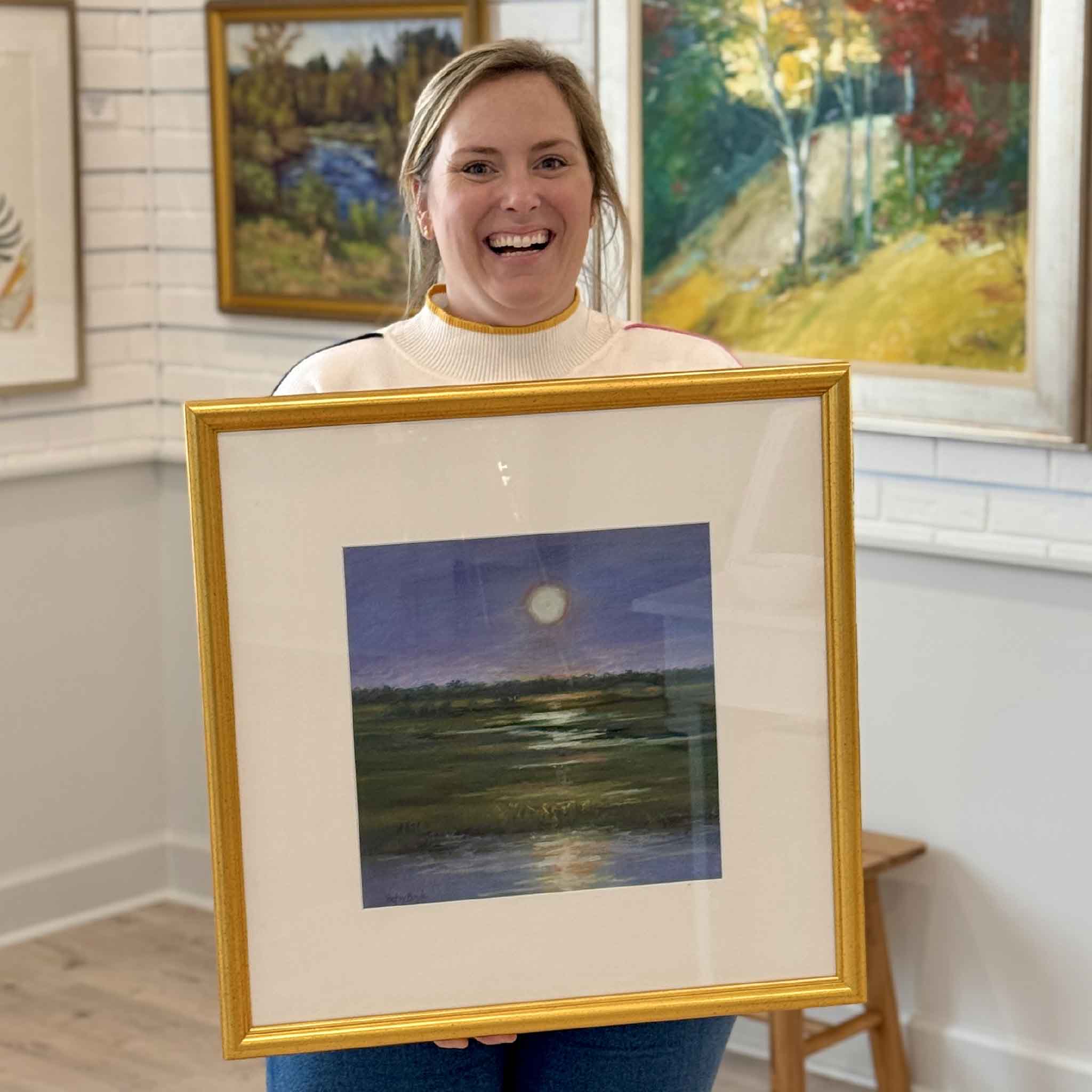 Owner of Gray Man Gallery holding “Moonlight Majesty,” an original pastel by Betsy Boyle depicting moonlight over a South Carolina Lowcountry marsh, professionally framed and displayed in Pawleys Island art gallery