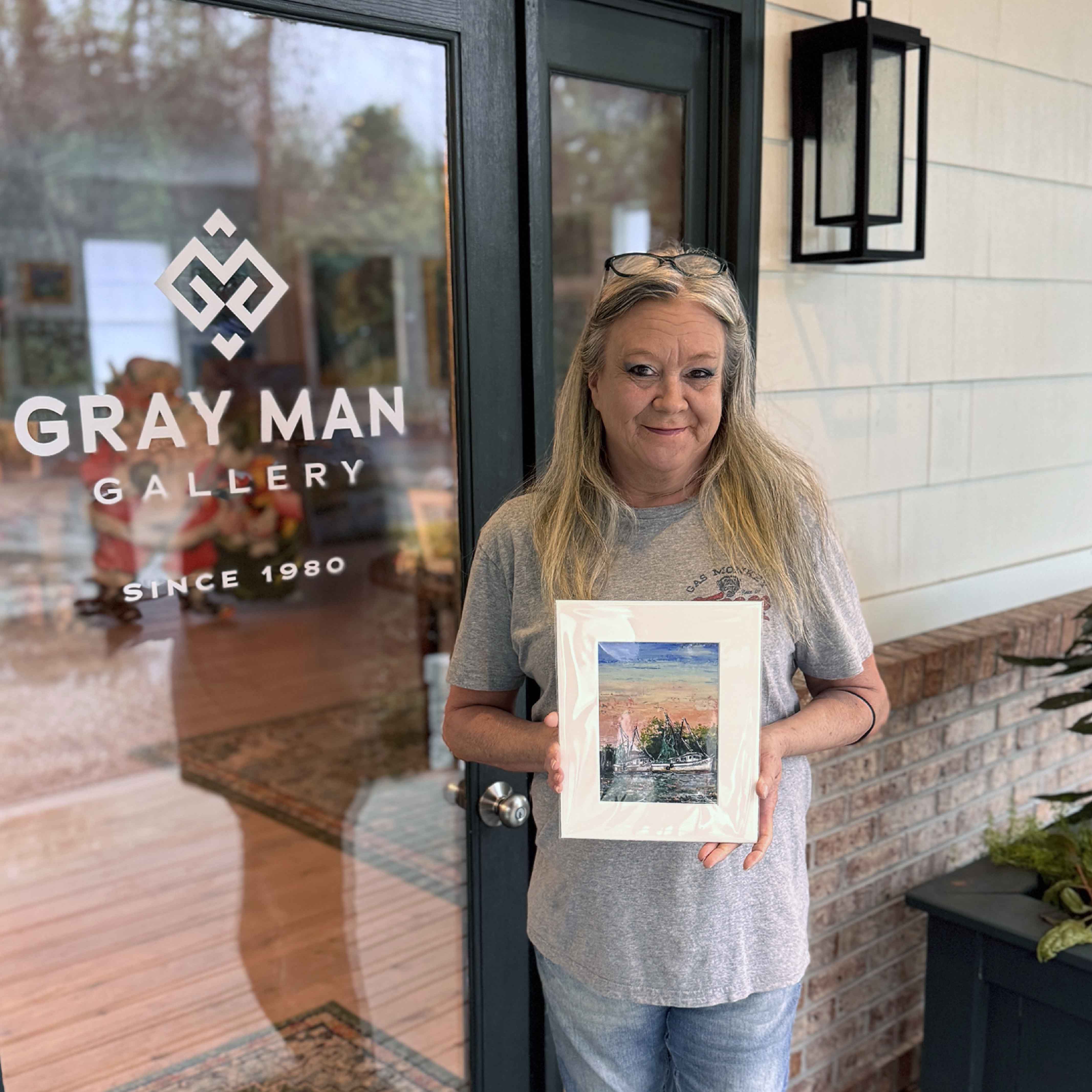 Charlotte holding Morning Harbor print by Jeanne O’Neal displayed in a white mat against a white brick wall, featuring shrimp boats resting beneath a soft coral and blue sunrise. Pictured at Gray Man Gallery.