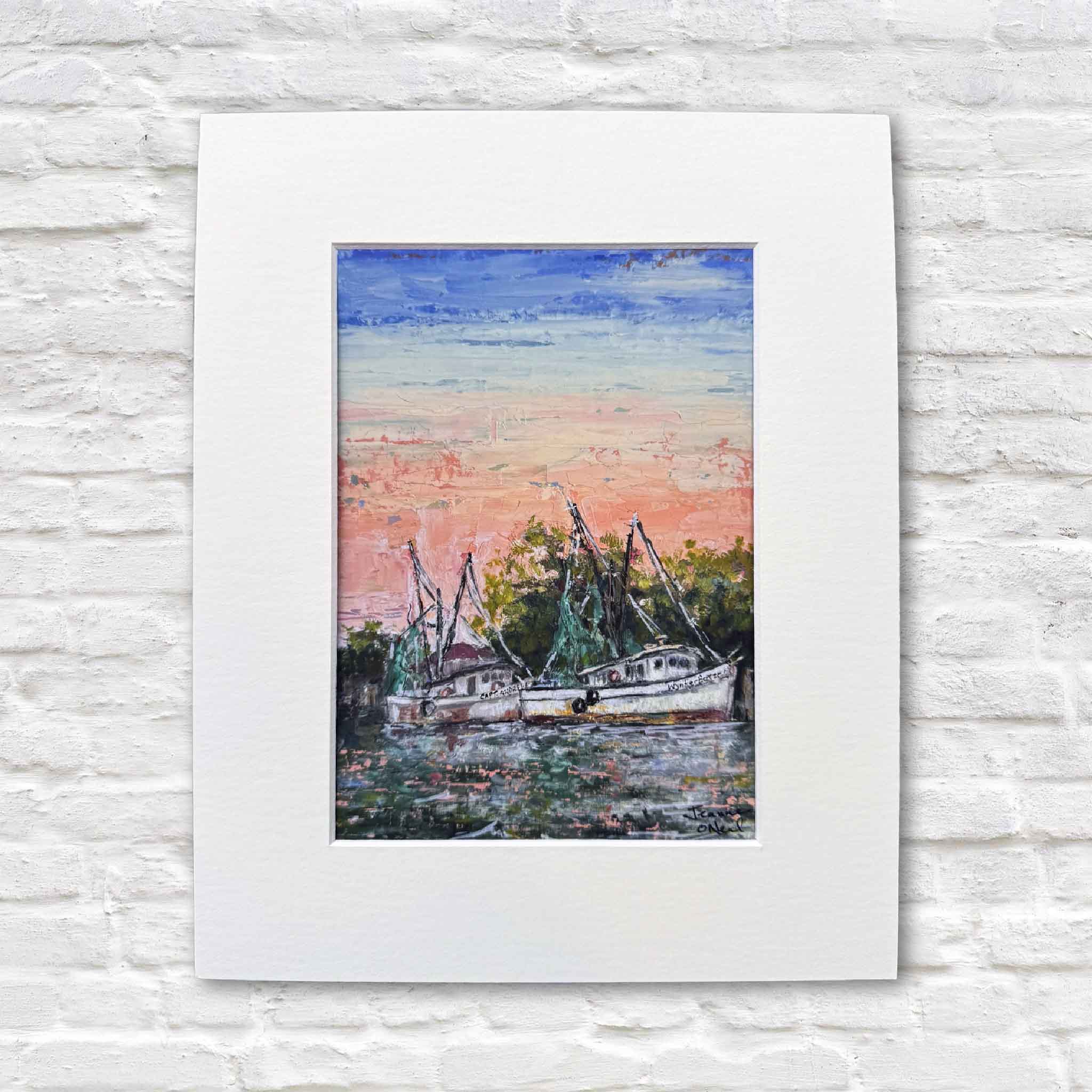 This photo shows the Morning Harbor print by Jeanne O’Neal displayed in a white mat against a white brick wall, featuring shrimp boats resting beneath a soft coral and blue sunrise.