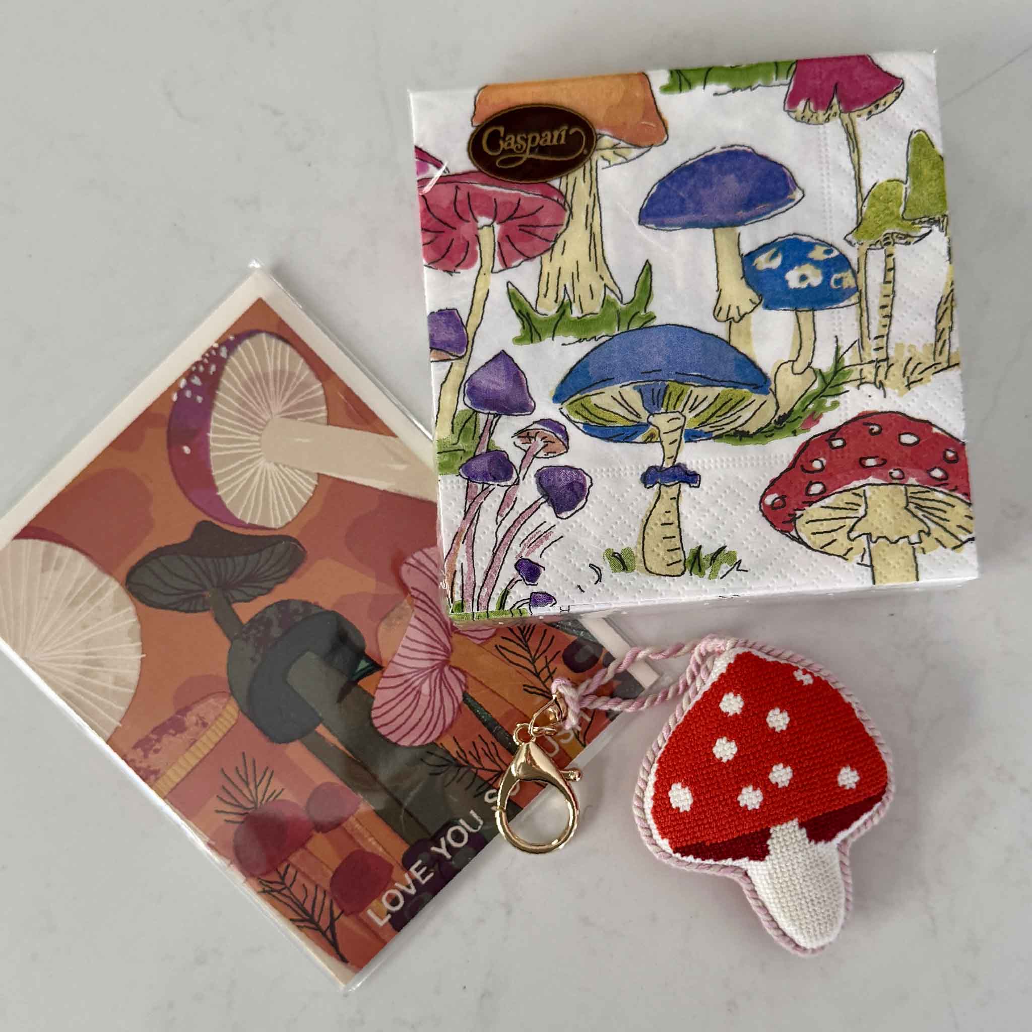 A whimsical mushroom-themed gift set featuring a hand-stitched red and white mushroom needlepoint bag charm, colorful mushroom cocktail napkins, and a coordinating mushroom greeting card, beautifully styled together at Gray Man Gallery in Pawleys Island, South Carolina. This playful collection makes a thoughtful gift for mushroom lovers, cottagecore enthusiasts, gardeners, hikers, and anyone who appreciates nature-inspired accessories and stationery.