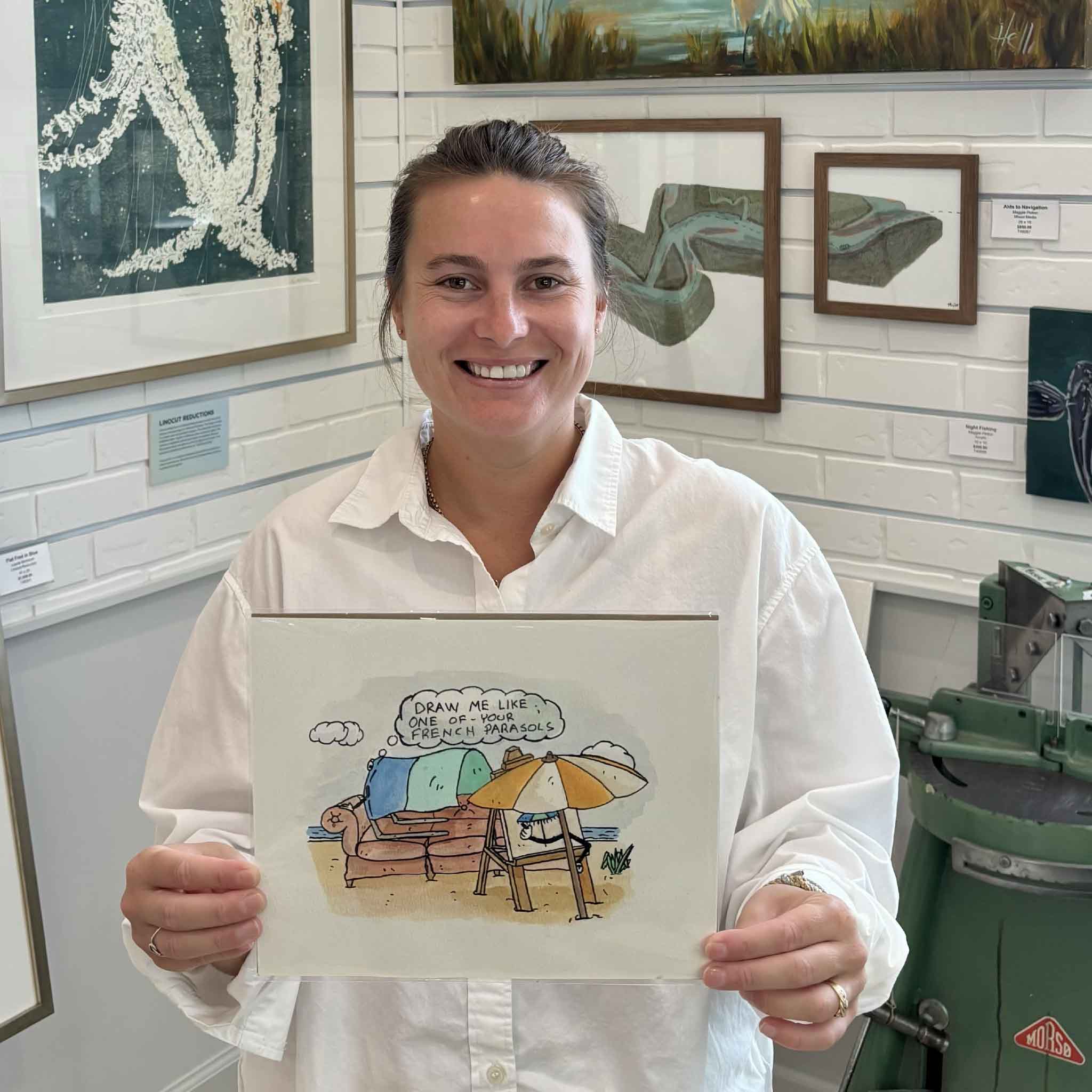 Kyra holding French Parasol + Shibumi by Charleston artist Nick kawczynski, an original pen and watercolor beach artwork featuring a humorous Titanic-inspired exchange between a Shibumi shade and traditional beach umbrella, inside Gray Man Gallery in Pawleys Island.