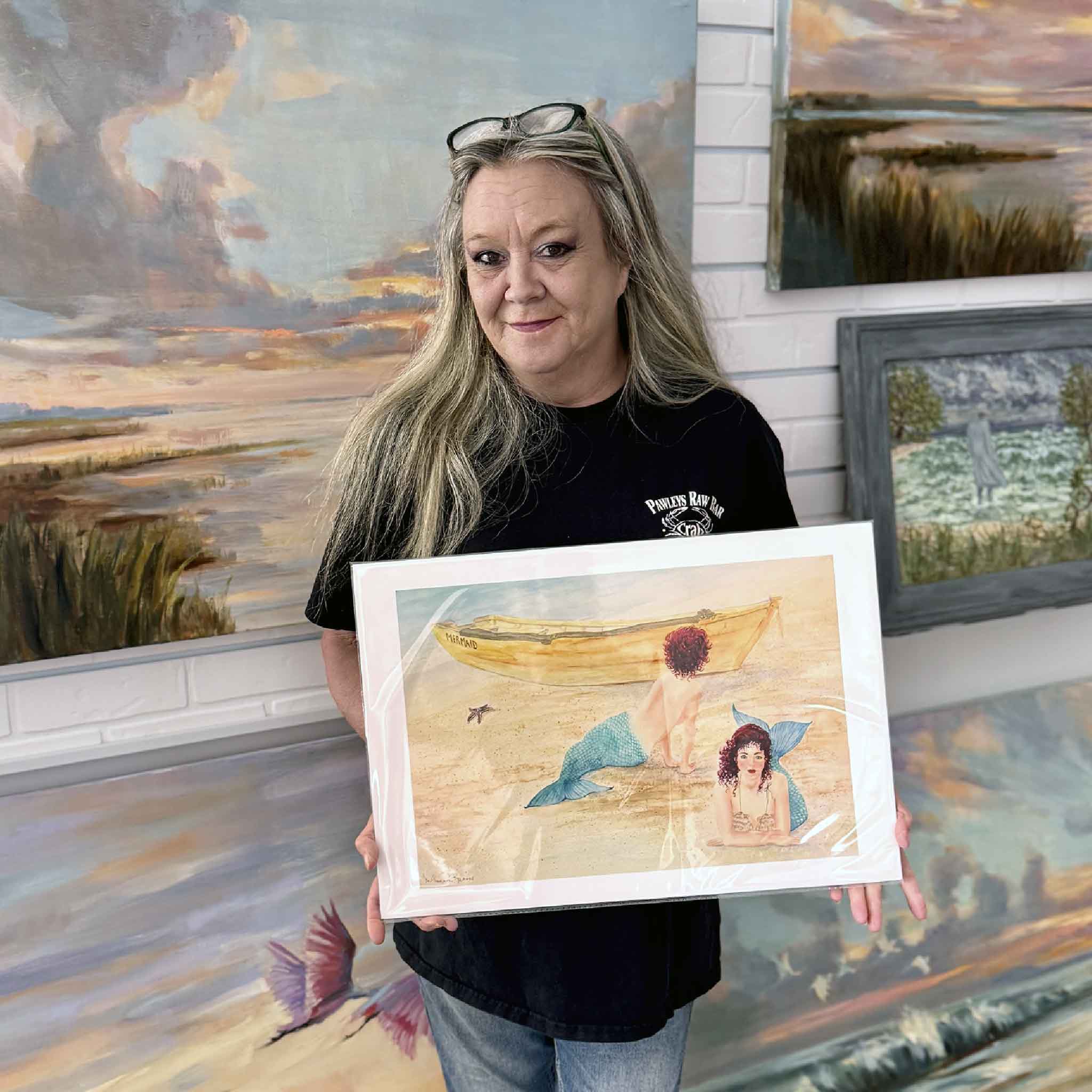 A Gray Man Gallery team member holds Our Name Must Be Ours by Mimi Beaver, an original watercolor depicting two mermaids resting on a sandy shore near a weathered yellow boat, donated to support Salty Dogs Senior Animal Rescue.