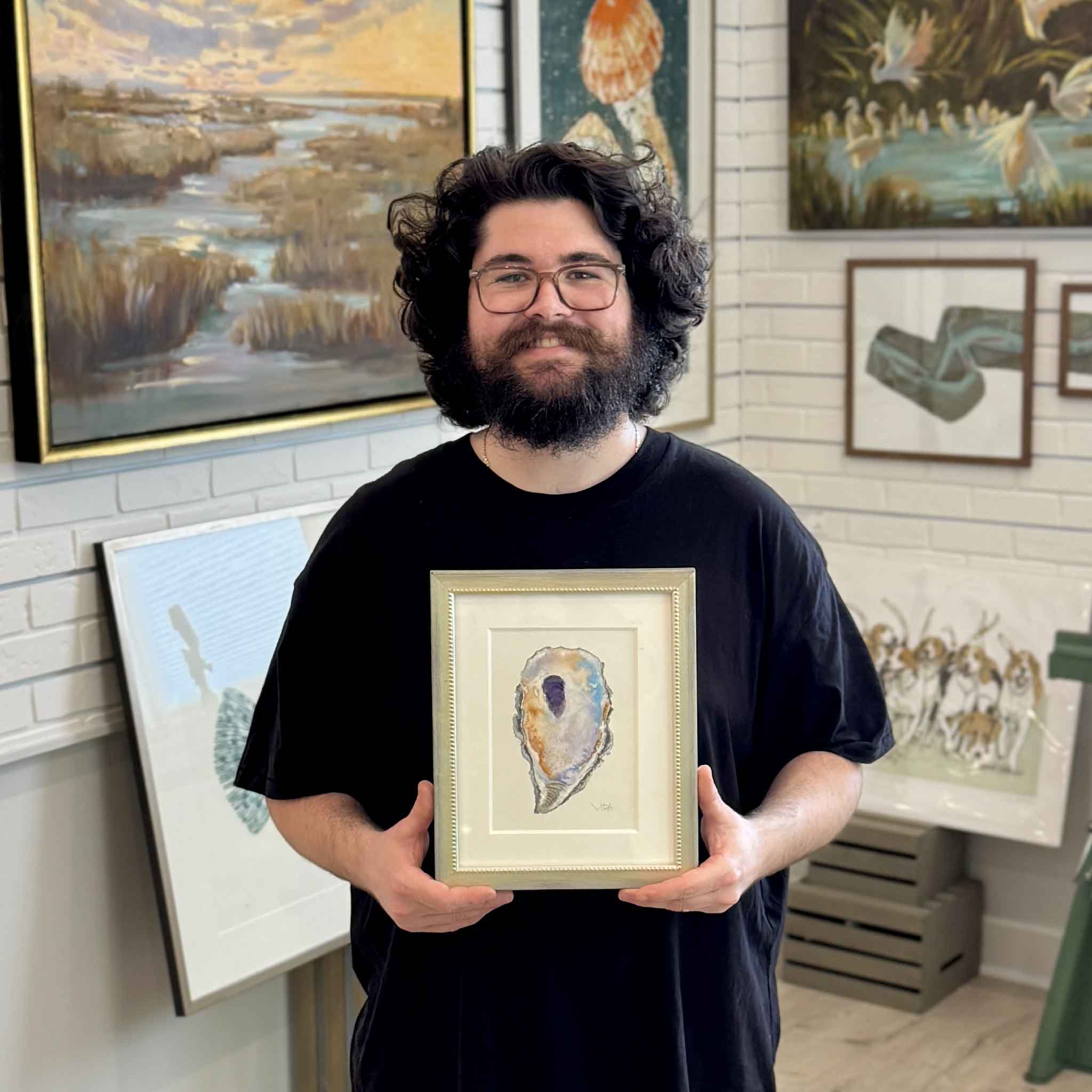Gray Man Gallery team member holding Pawleys Oyster VII, an original framed watercolor by Pawleys Island artist Vida Miller featuring a colorful Lowcountry oyster shell, shown inside Gray Man Gallery in Pawleys Island, South Carolina, with original coastal artwork displayed throughout the gallery.