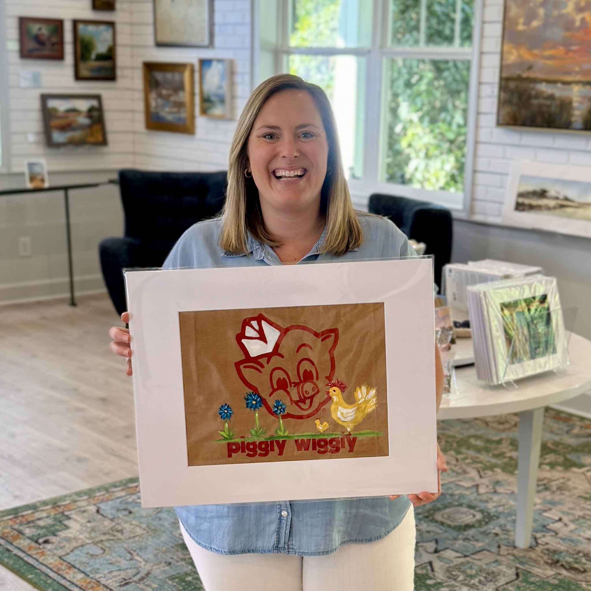 Gray Man Gallery team member holding Piggly Wiggly Mama Hen and Chick painting by local artist Jenny Reynolds, showing size and detail of grocery bag artwork