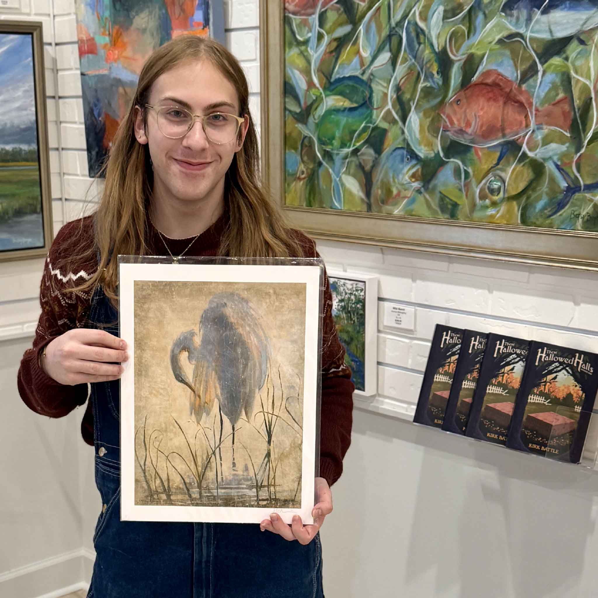 A gallery assistant holds the 12x16 Preening Egret art print by Helli Luck, showing the scale and elegant detail of the Lowcountry bird illustration against a backdrop of original coastal artwork at Gray Man Gallery.