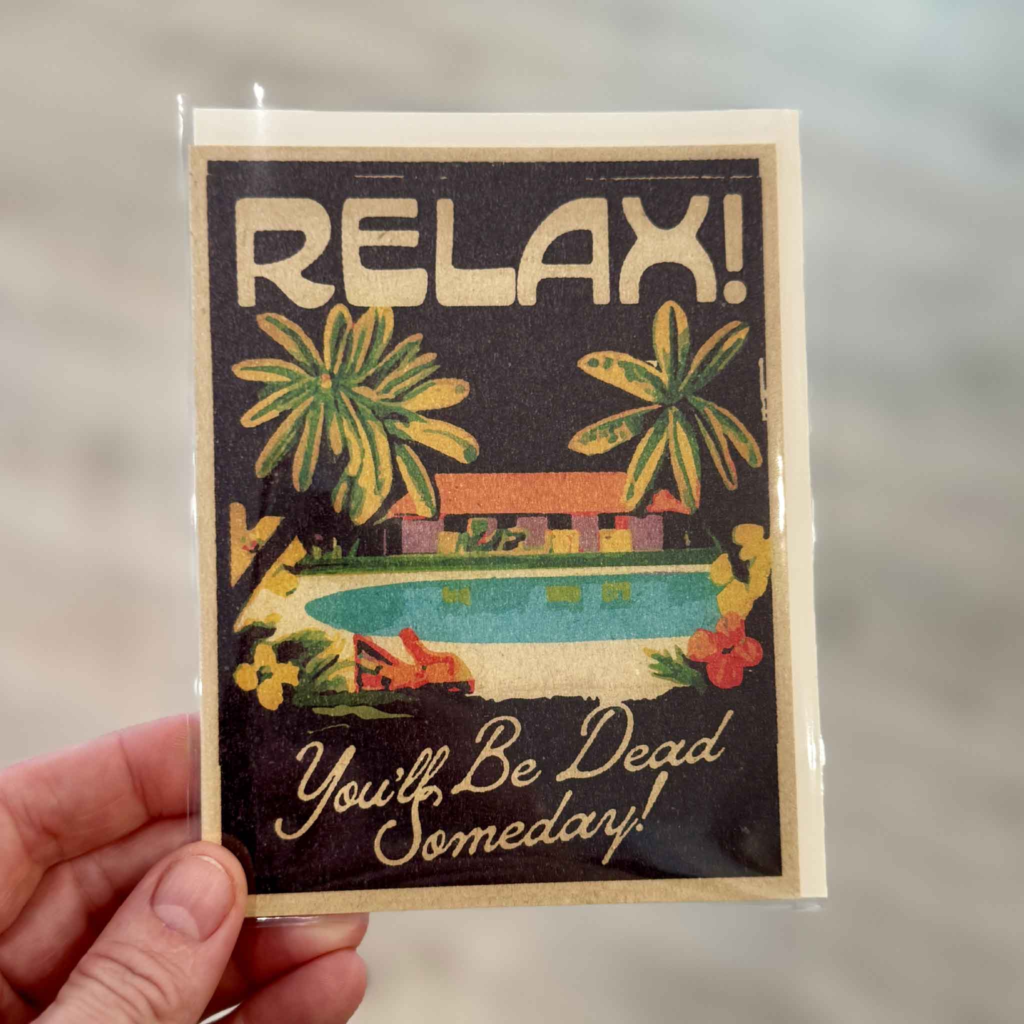 Hand holding the Red Cap Cards "Relax! You'll Be Dead Someday" greeting card illustrated by Daren Thomas Magee inside Gray Man Gallery in Pawleys Island, South Carolina. Funny blank-inside greeting card featuring a retro tropical pool scene with palm trees and playful lettering.