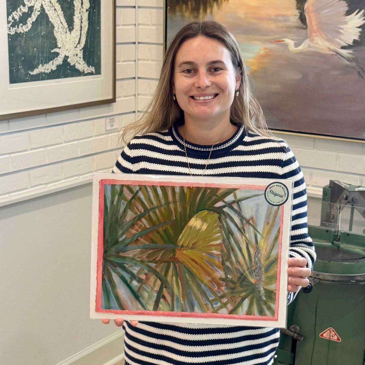 Gray Man Gallery team member holding Palmetto Fronds #212, an original oil on paper by local artist Retta Dotterer, showing the scale, layered palmetto foliage and hand-painted pink border inside the Pawleys Island gallery.