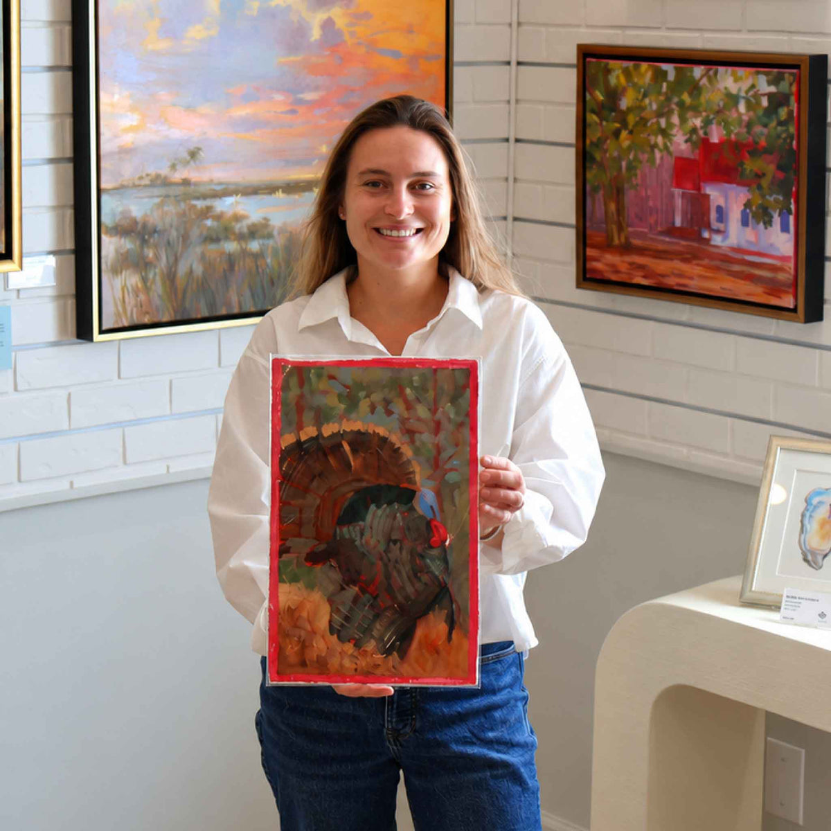 Gray Man Gallery team member holding Turkey Solo #210, an original wild turkey oil on paper by local artist Retta Dotterer, showing the artwork’s scale and hand-painted pink border inside the Pawleys Island gallery.