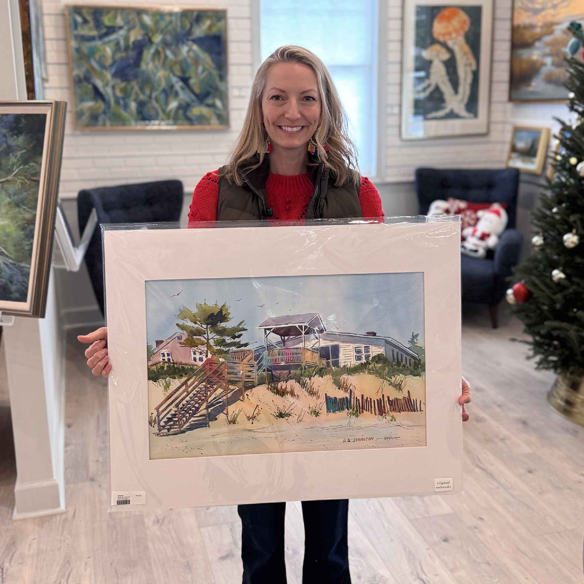 Local artist Jeanne O'Neal holds Gary Johnston’s matted Rowell’s Retreat watercolor, showing the beach house scene and its detailed coastal brushwork.